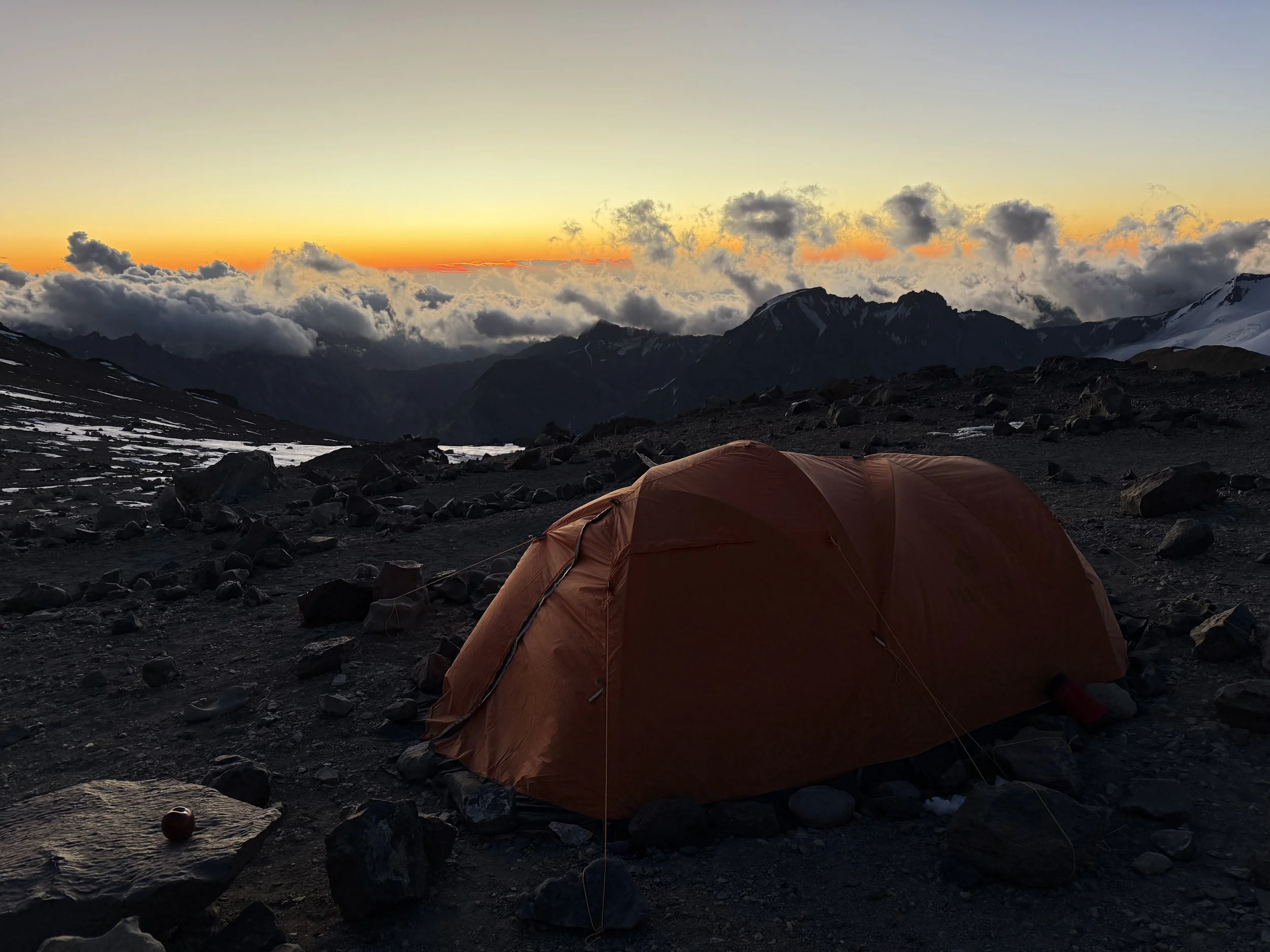 A tent set up on rocky terrain with snow patches, overlooking mountain peaks and clouds at sunset.