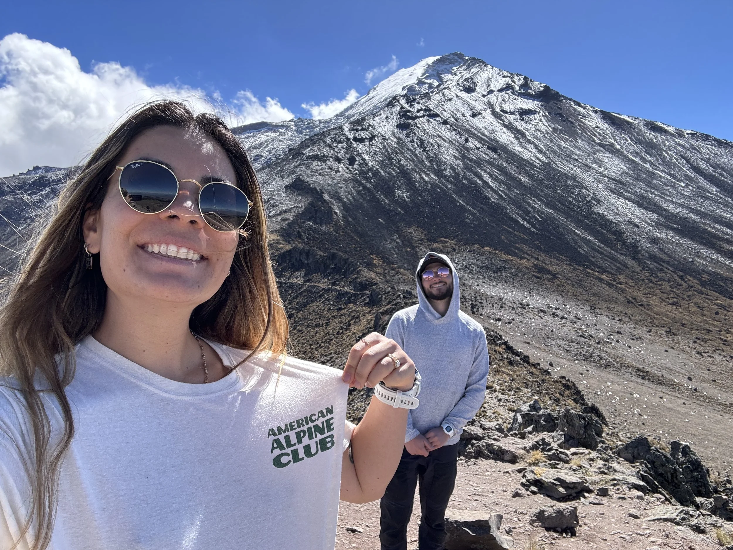 Two people outdoors in a mountainous area with snow-covered peaks. The woman in the foreground is smiling, wearing sunglasses and a white shirt with "American Alpine Club" written on it. The man in the background is standing on rocky terrain, wearing