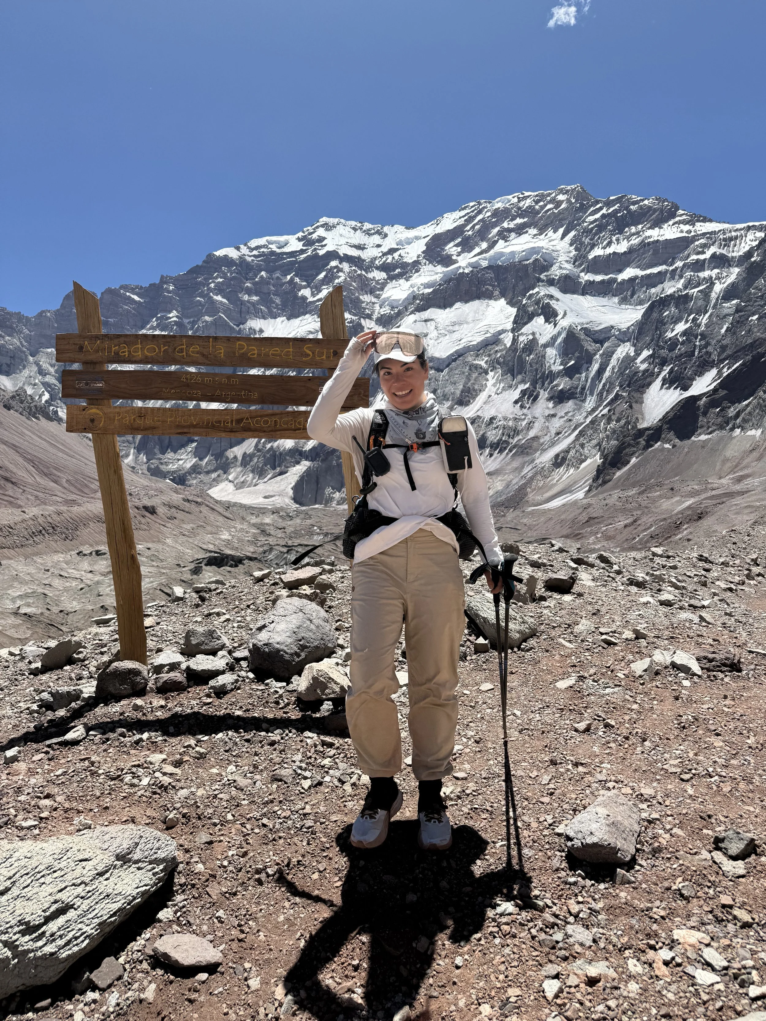 A woman smiling and dressed in hiking gear standing on rocky terrain at a high altitude, holding hiking poles, with snow-covered mountains and a blue sky in the background. She is near a wooden sign that reads 'Mirador de la Pared Sur,' indicating sh