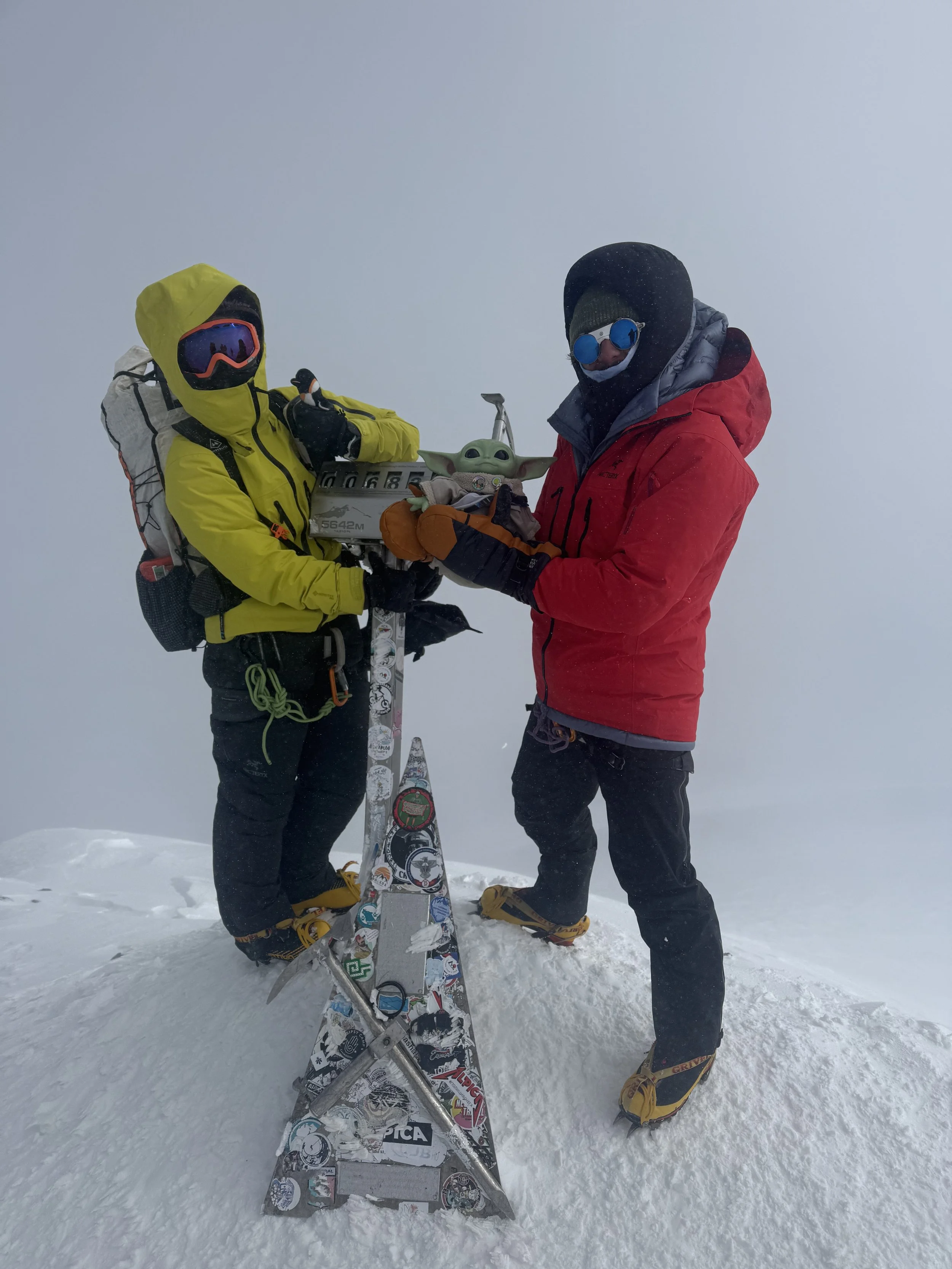 Two mountaineers in winter gear standing on snow-covered peak with a carved Baby Yoda figure.
