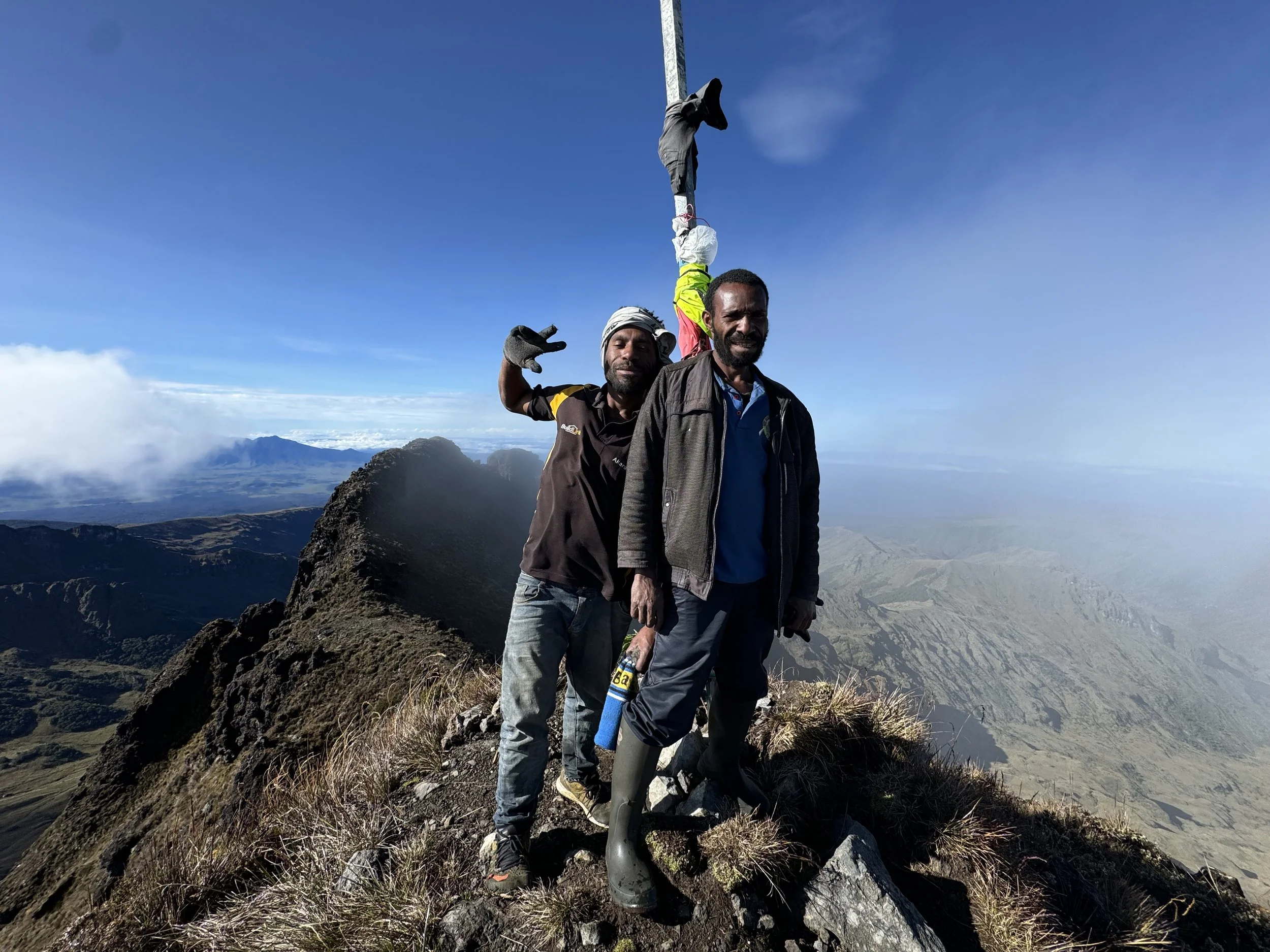 Three men standing on a mountain summit with scenic landscape in background, one making a peace sign, some clouds, and a partly sunny sky.