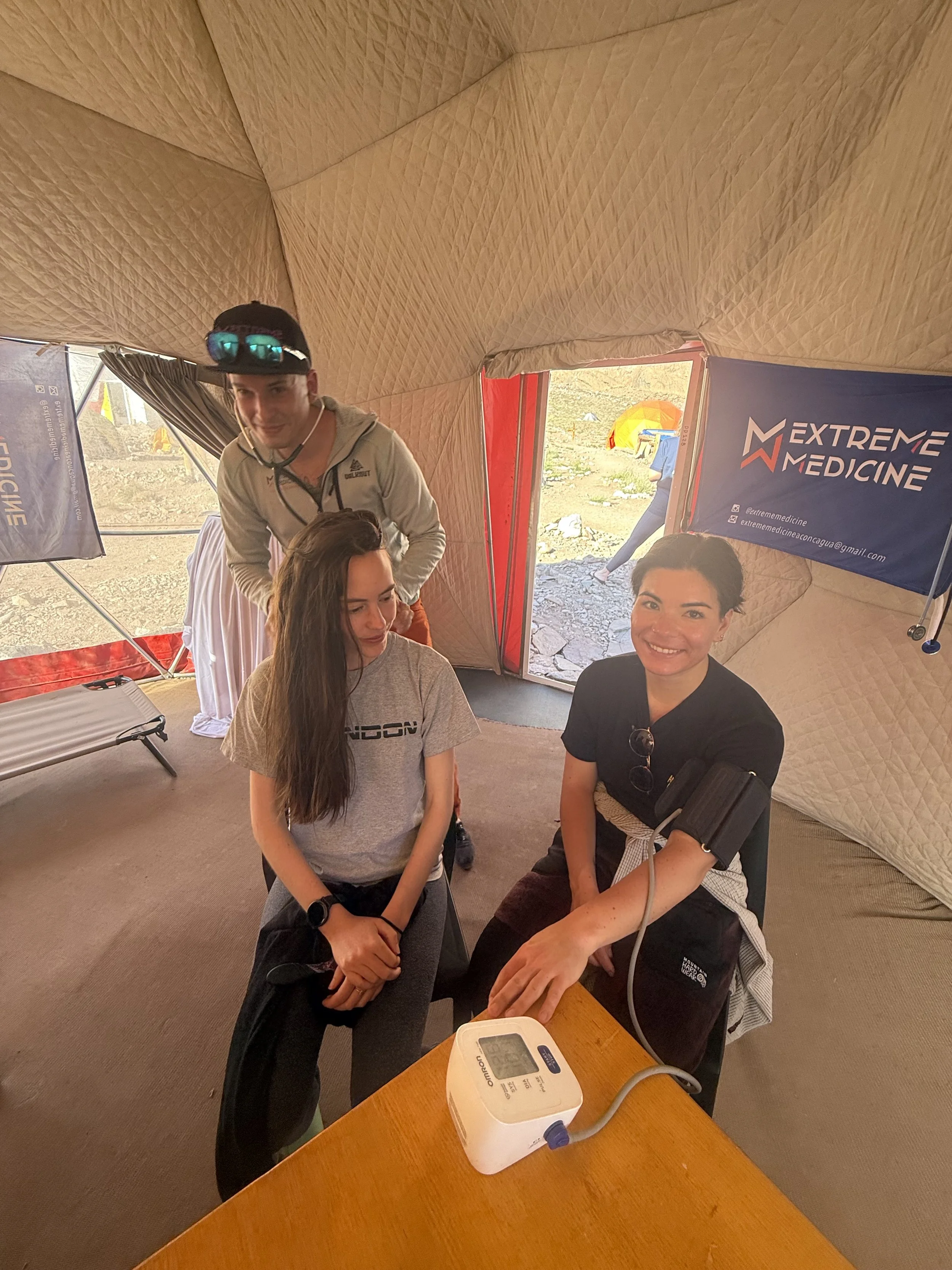 A young woman has her blood pressure checked by a medical worker inside a beige tent with a banner that reads Extreme Medicine. Two other young people are nearby, one sitting and one standing, smiling and observing.