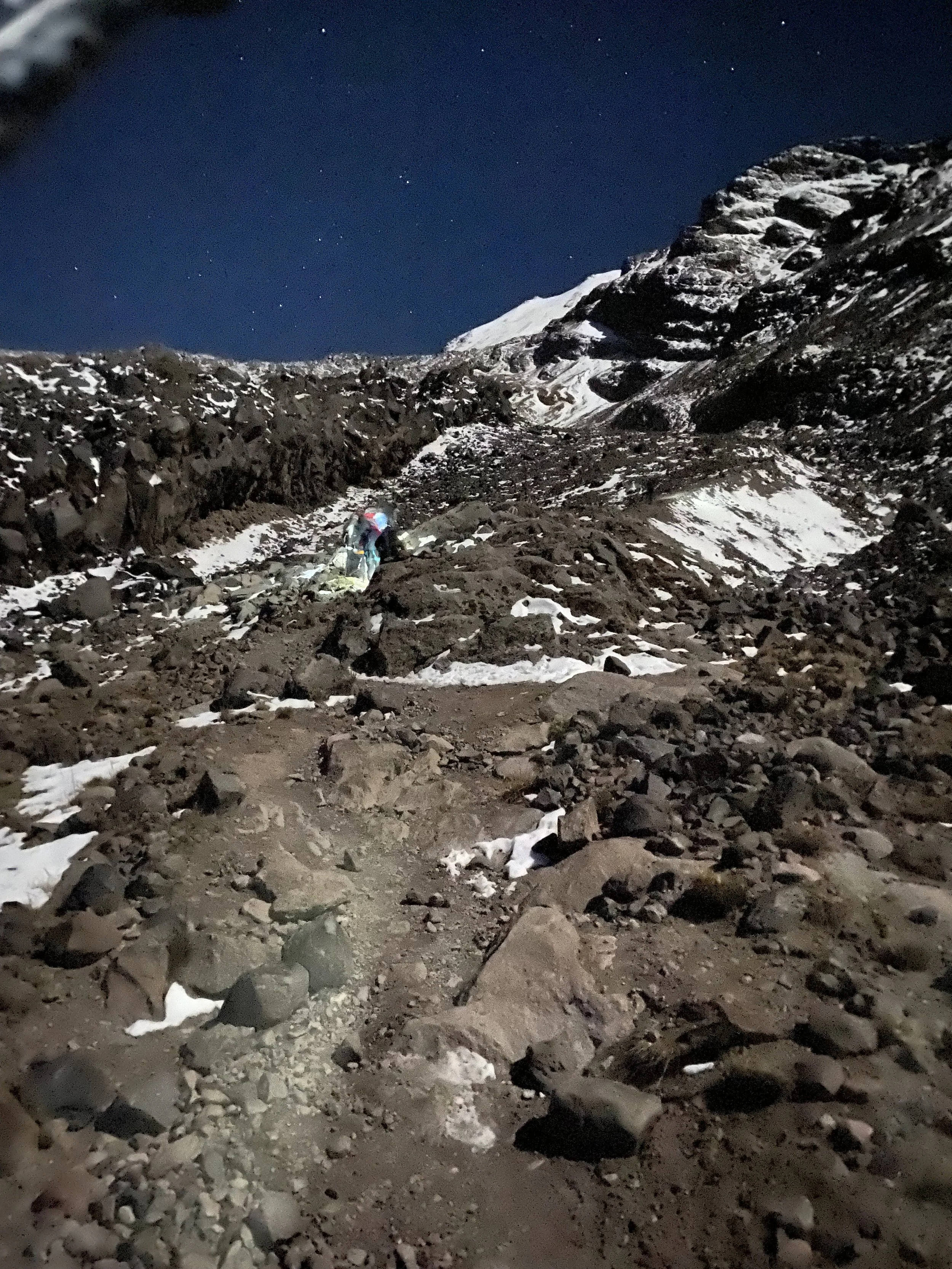 A rocky mountain trail at night with snow patches and a person wearing a helmet and headlamp hiking.