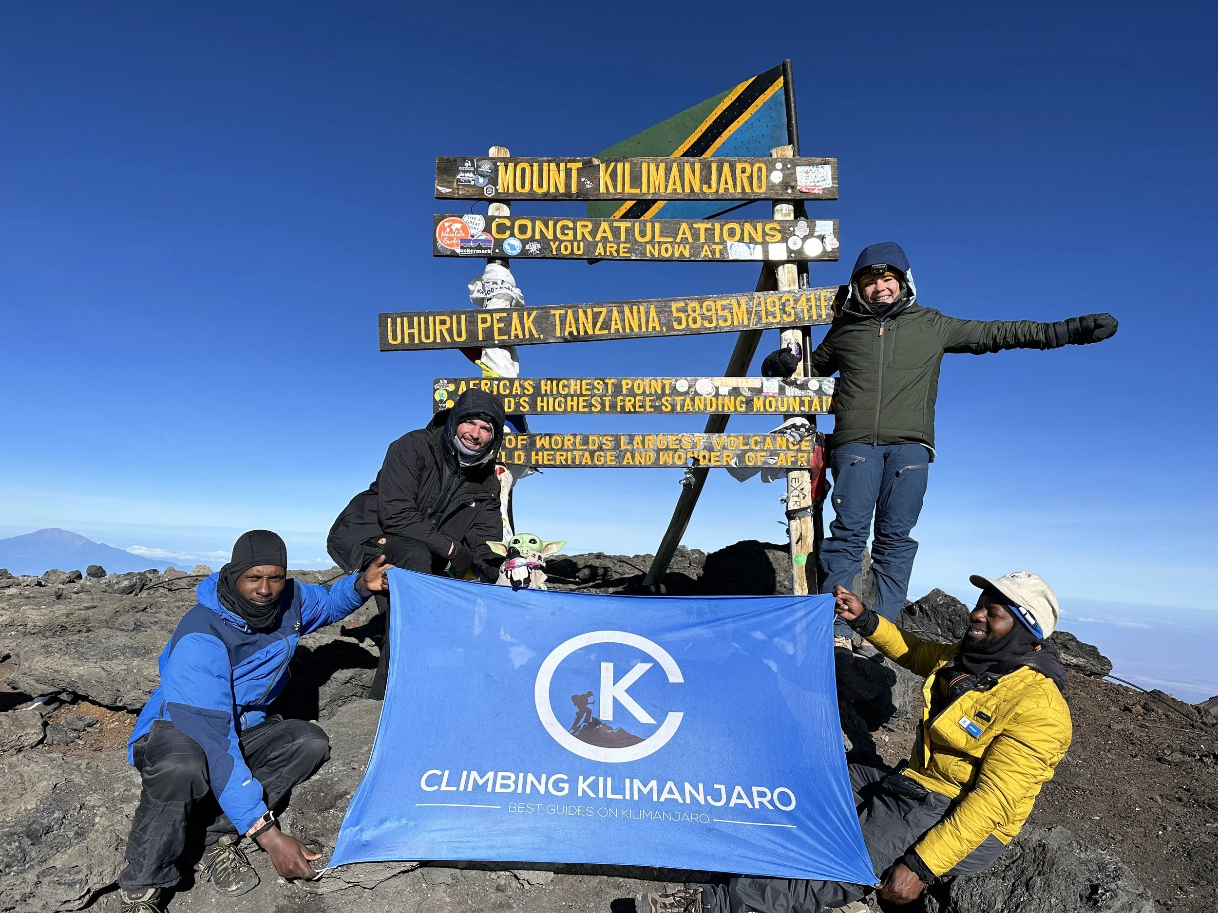 Group of climbers holding a blue flag at Uhuru Peak, Mount Kilimanjaro, Tanzania, with a colorful sign behind them displaying information about the summit, including its elevation of 5895 meters and its status as Africa's highest point.