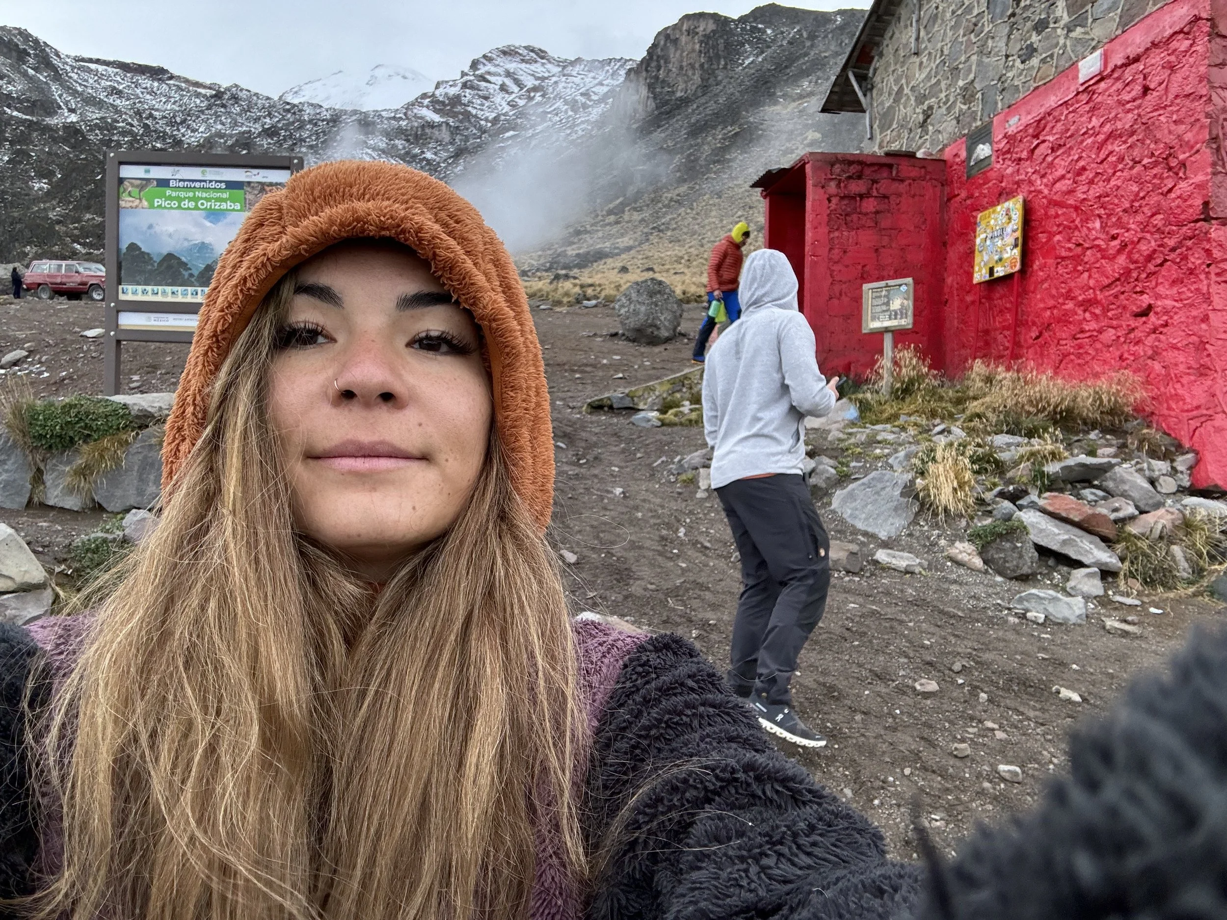 A woman wearing an orange hooded jacket taking a selfie at Machu Picchu with snowy mountains and a red building in the background.
