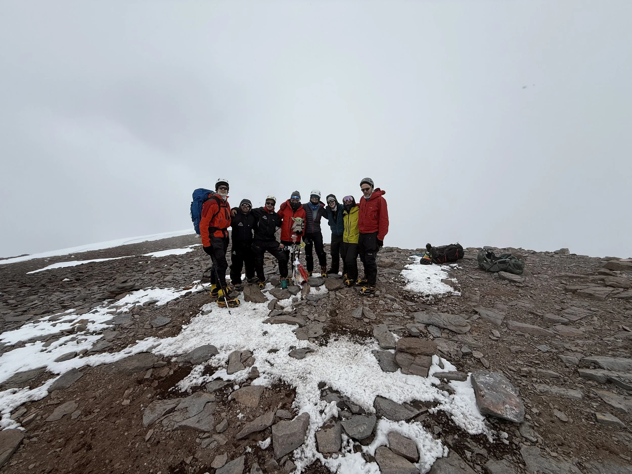 Group of hikers standing on a snowy mountain summit with backpacks and climbing gear, wearing helmets and outdoor jackets.