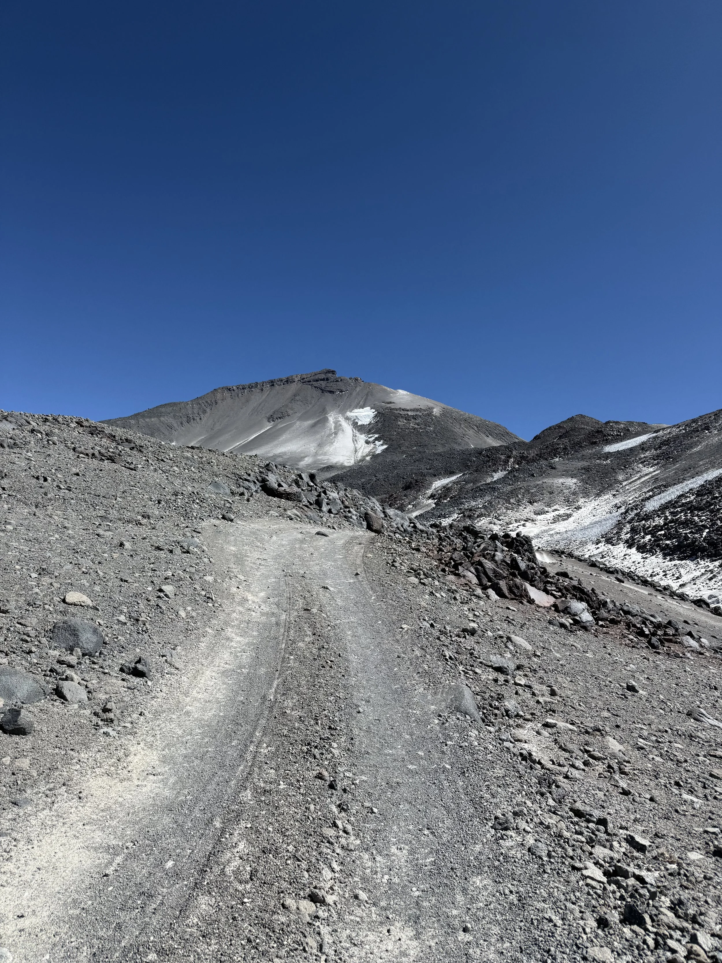 A rugged dirt mountain road with loose rocks, leading towards snow-capped peaks under a clear blue sky.