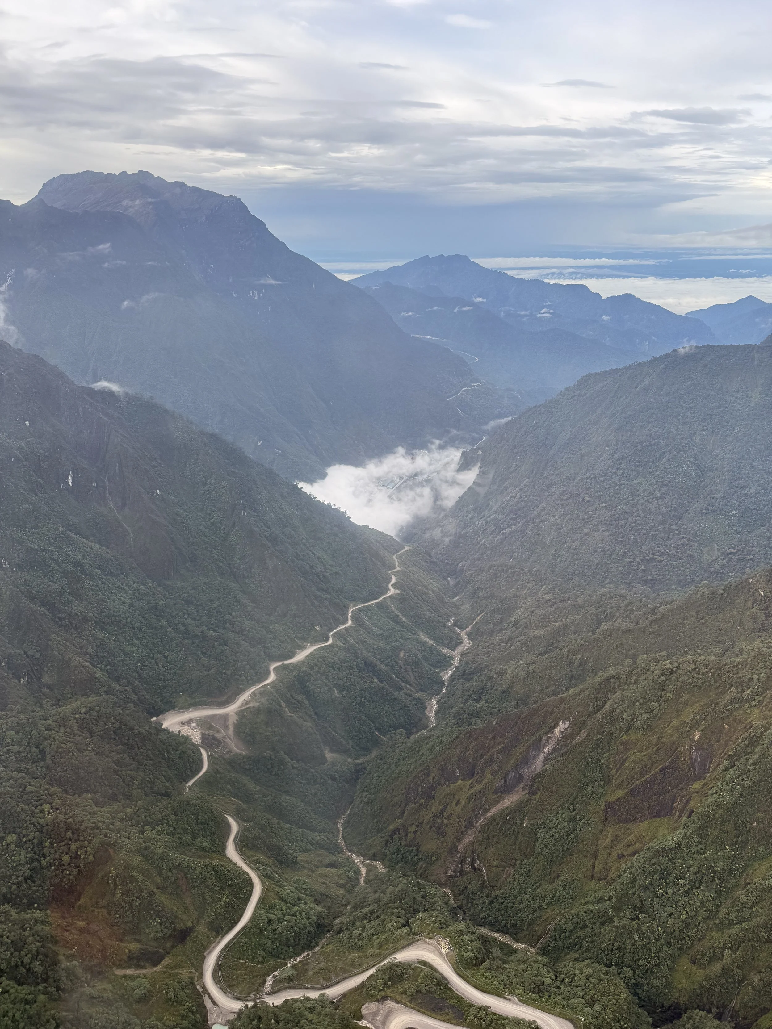 Mountain landscape with winding dirt roads and cloud-covered peaks under an overcast sky.