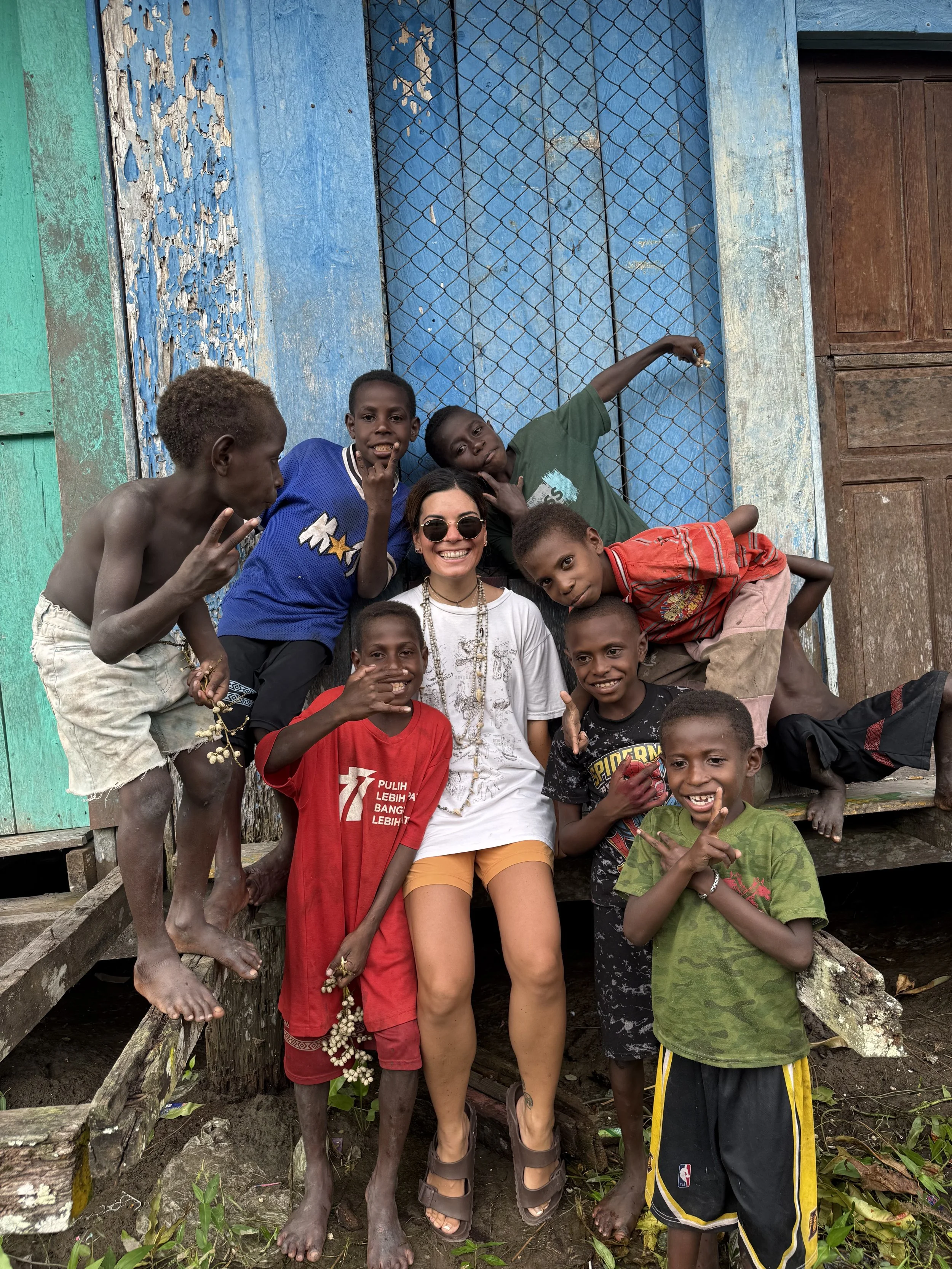 A woman with sunglasses and white clothing surrounded by children of various ages outside, standing in front of a rustic blue and green wooden structure with a metal gate.