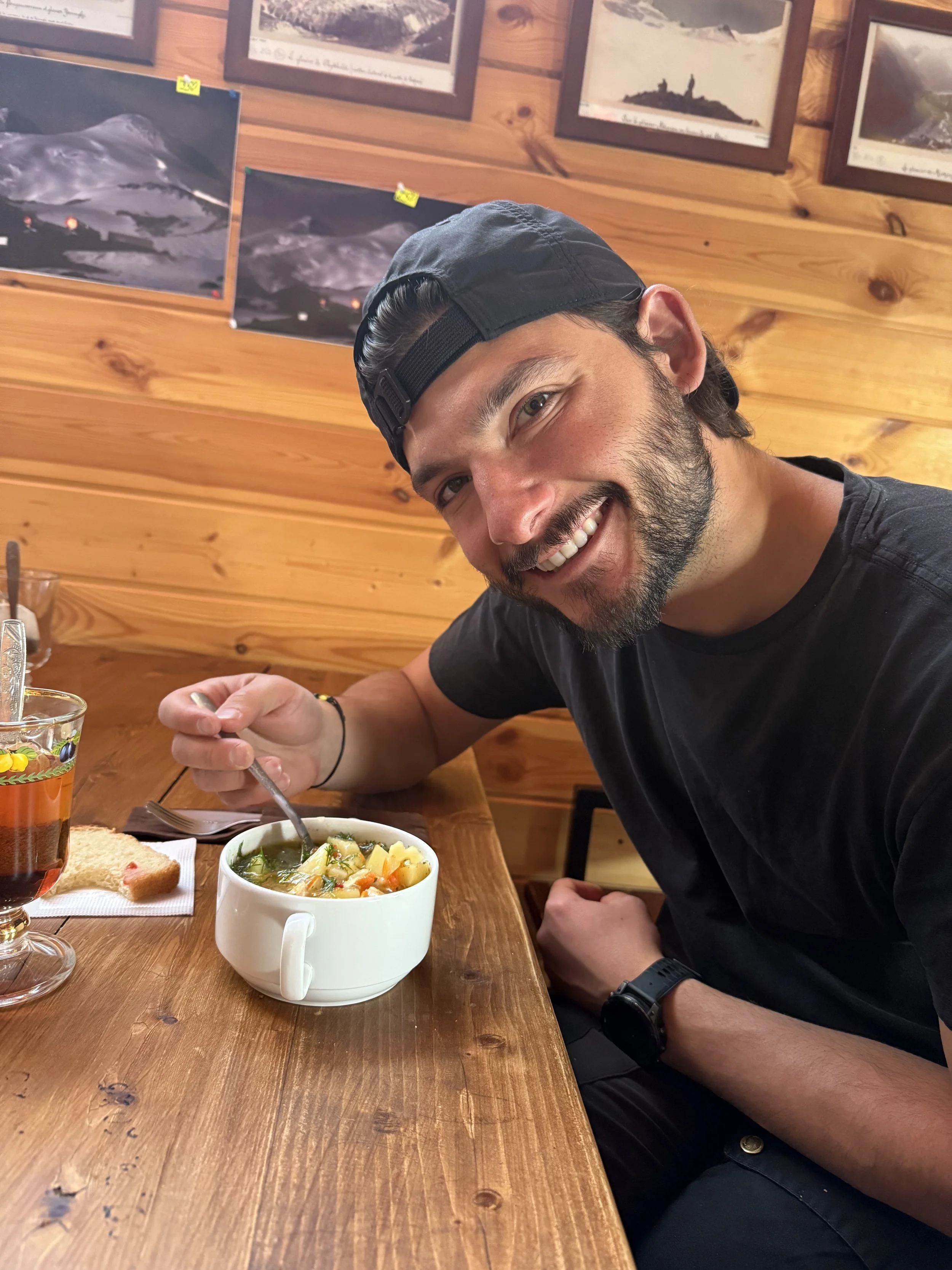 A man with a beard and a black cap smiling at the camera while eating a bowl of vegetable soup in a wooden restaurant. There is a glass of iced tea and a piece of bread on the table.