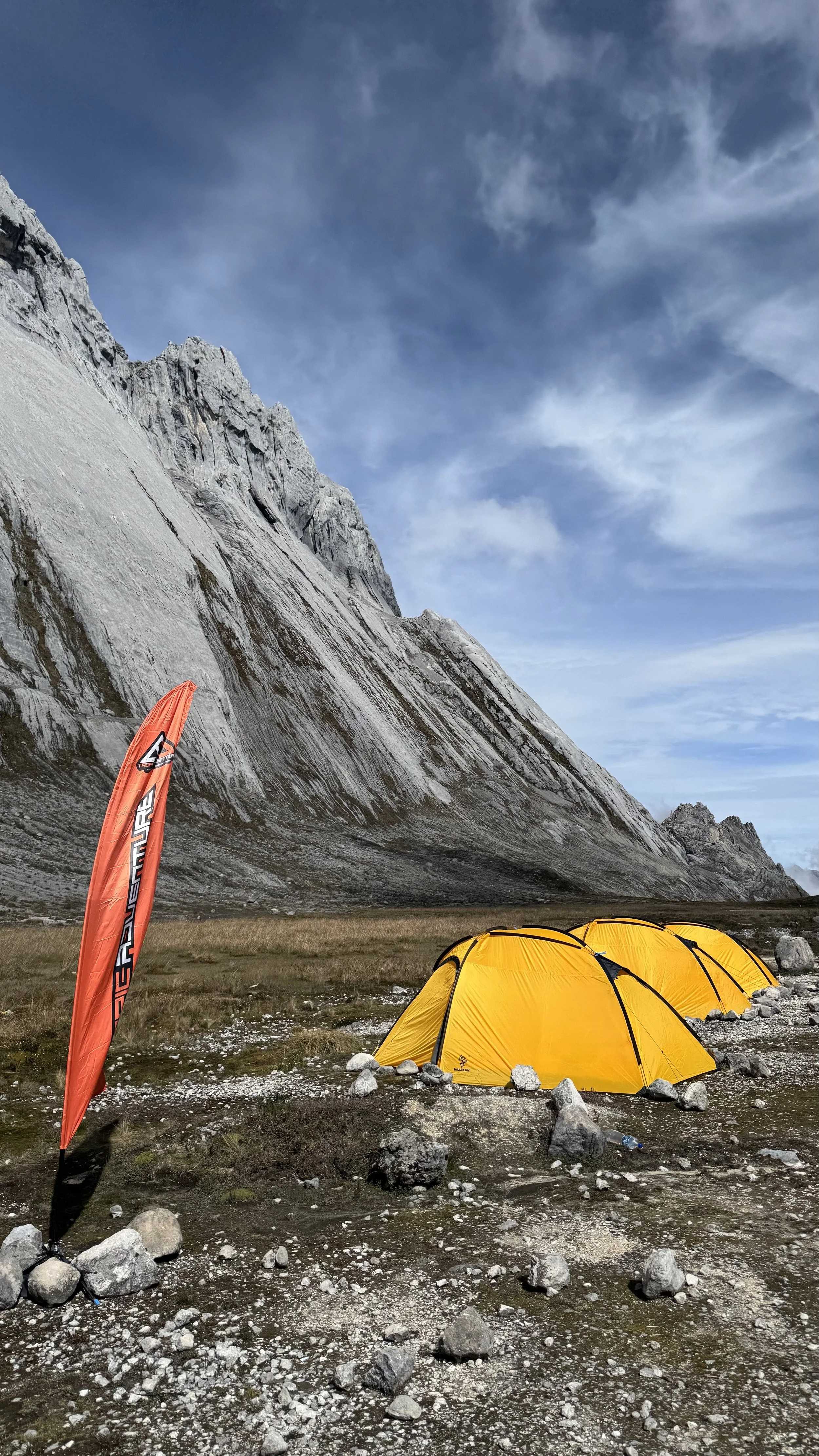 Three yellow camping tents pitched in a rocky area at the base of a large mountain with steep, jagged rock formations and a partly cloudy sky.