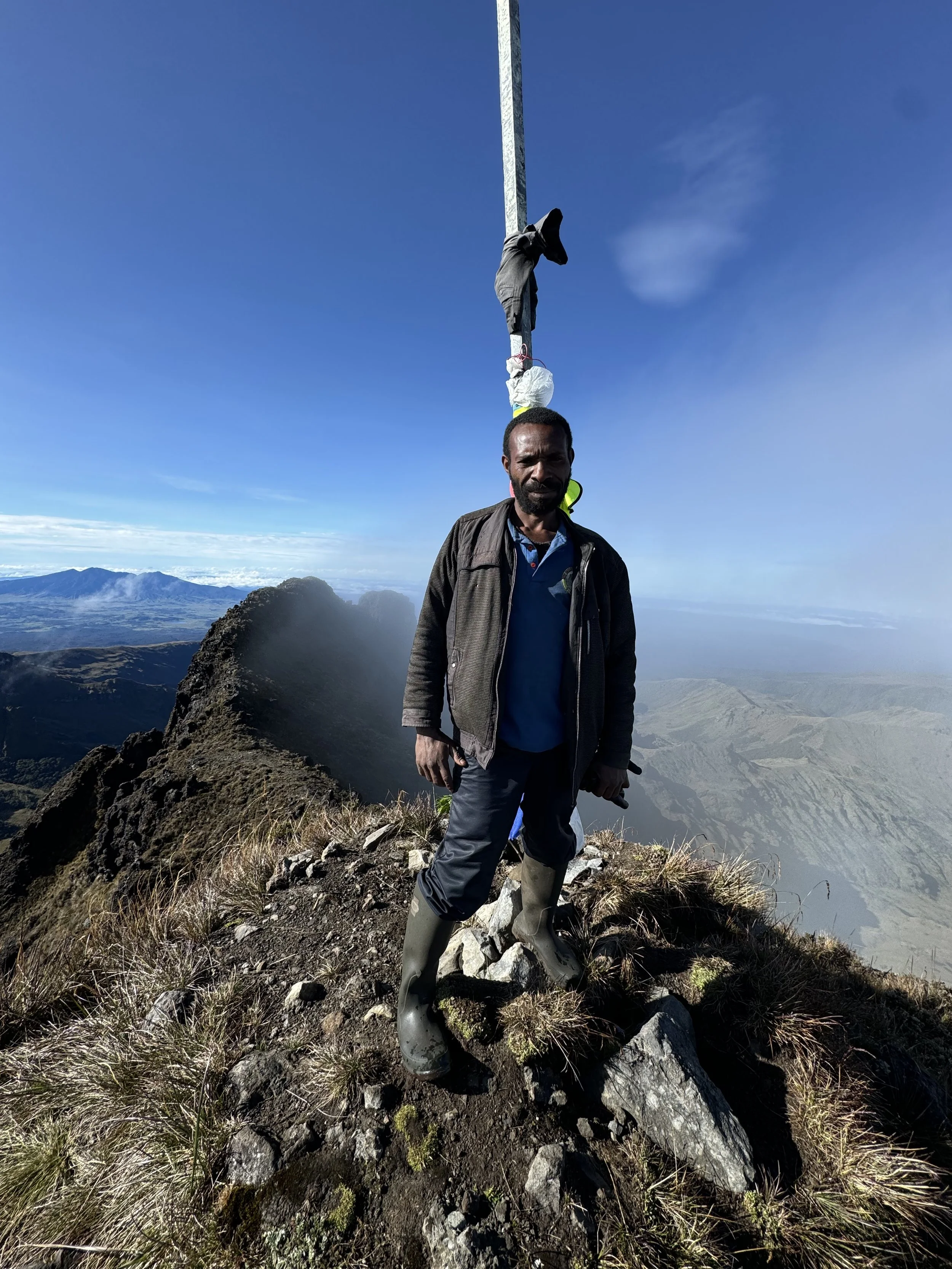 A man standing on a mountain summit with a tall pole behind him, wearing boots, with a scenic landscape and blue sky in the background.