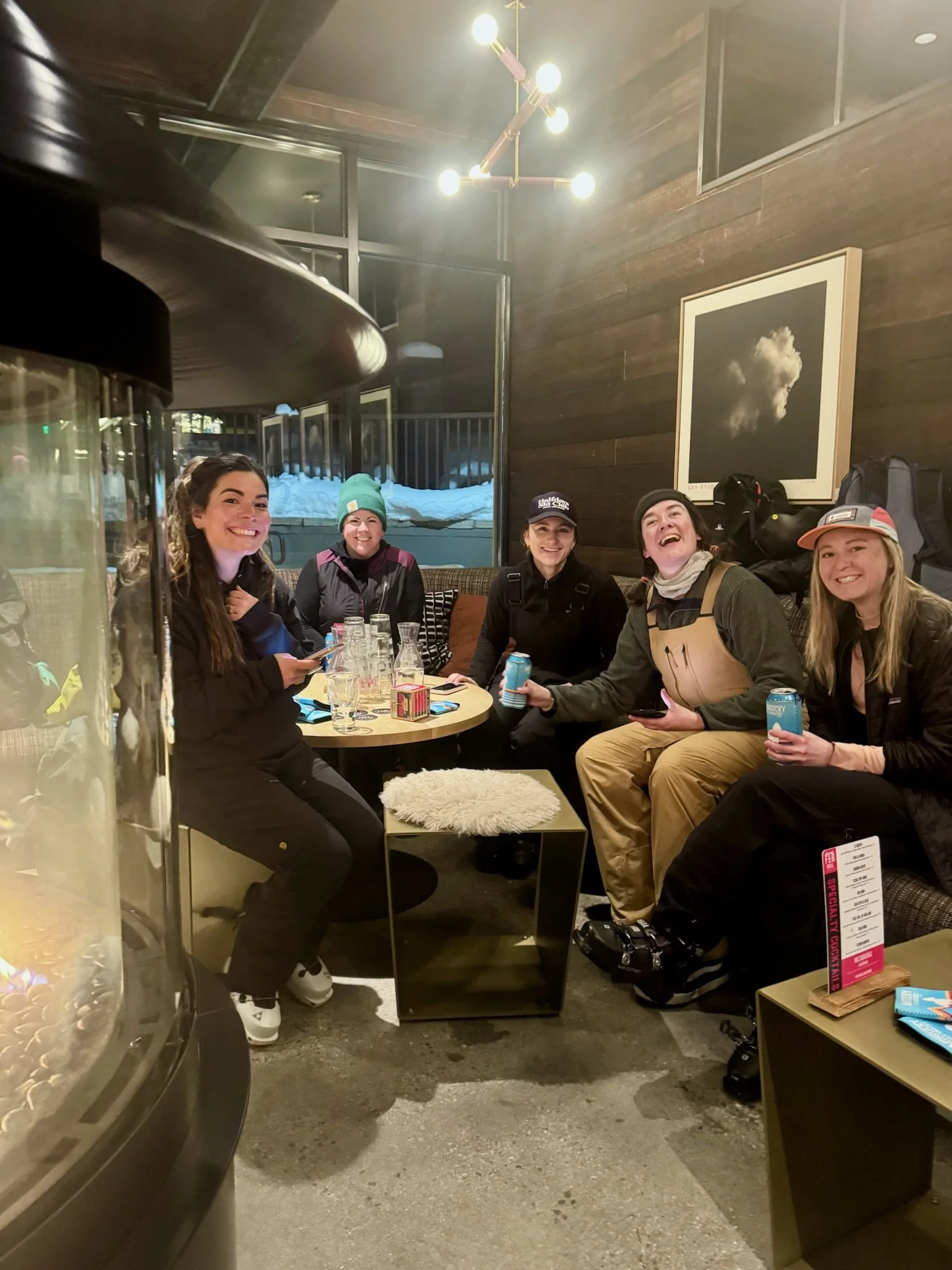 Five women sitting together at a table in a cozy, wooden-walled cafe or lounge, smiling and holding drinks, with snow outside the window.