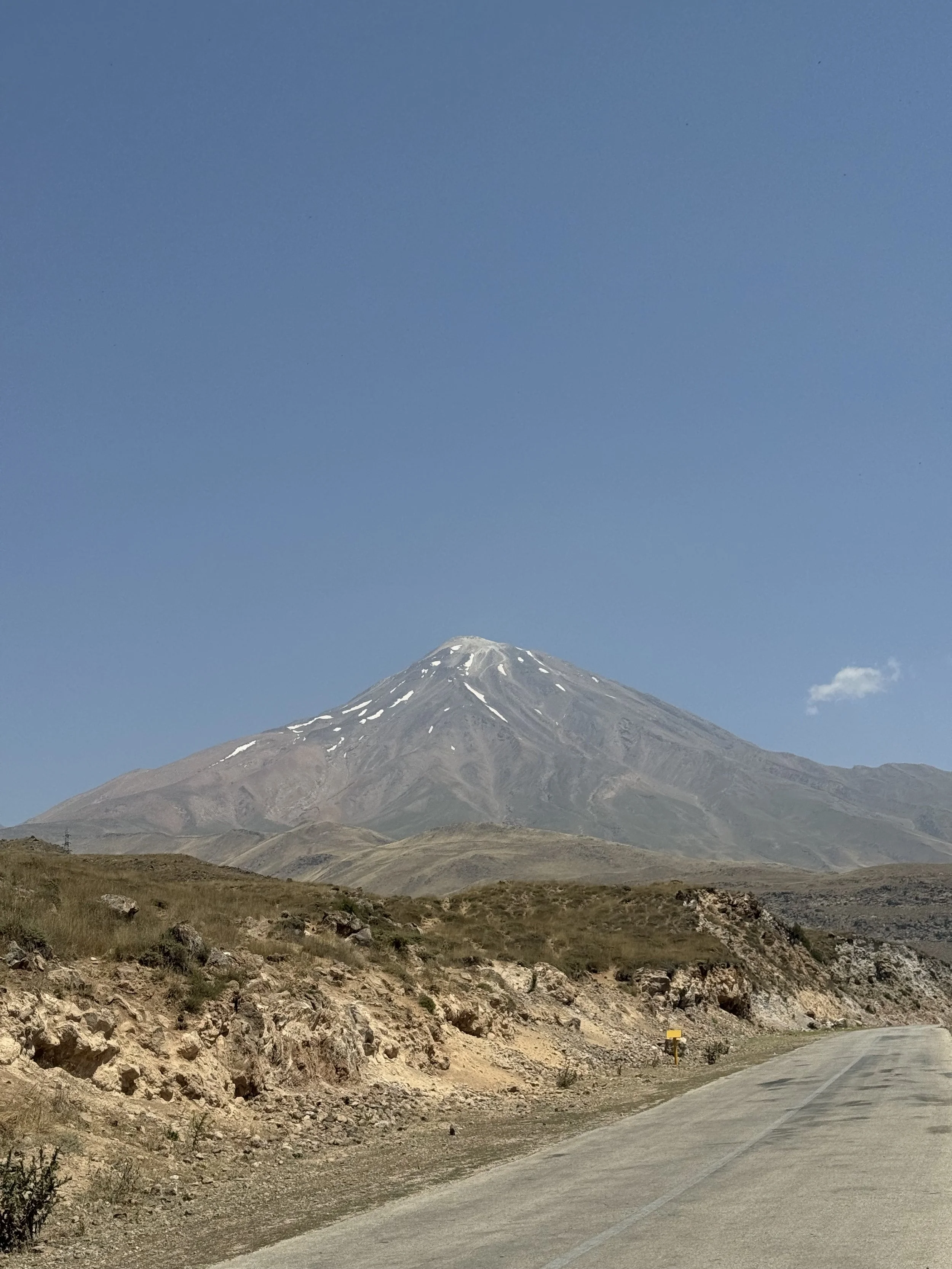 A mountain with a snow cap, under a clear blue sky, near a paved road in a dry, rocky landscape.