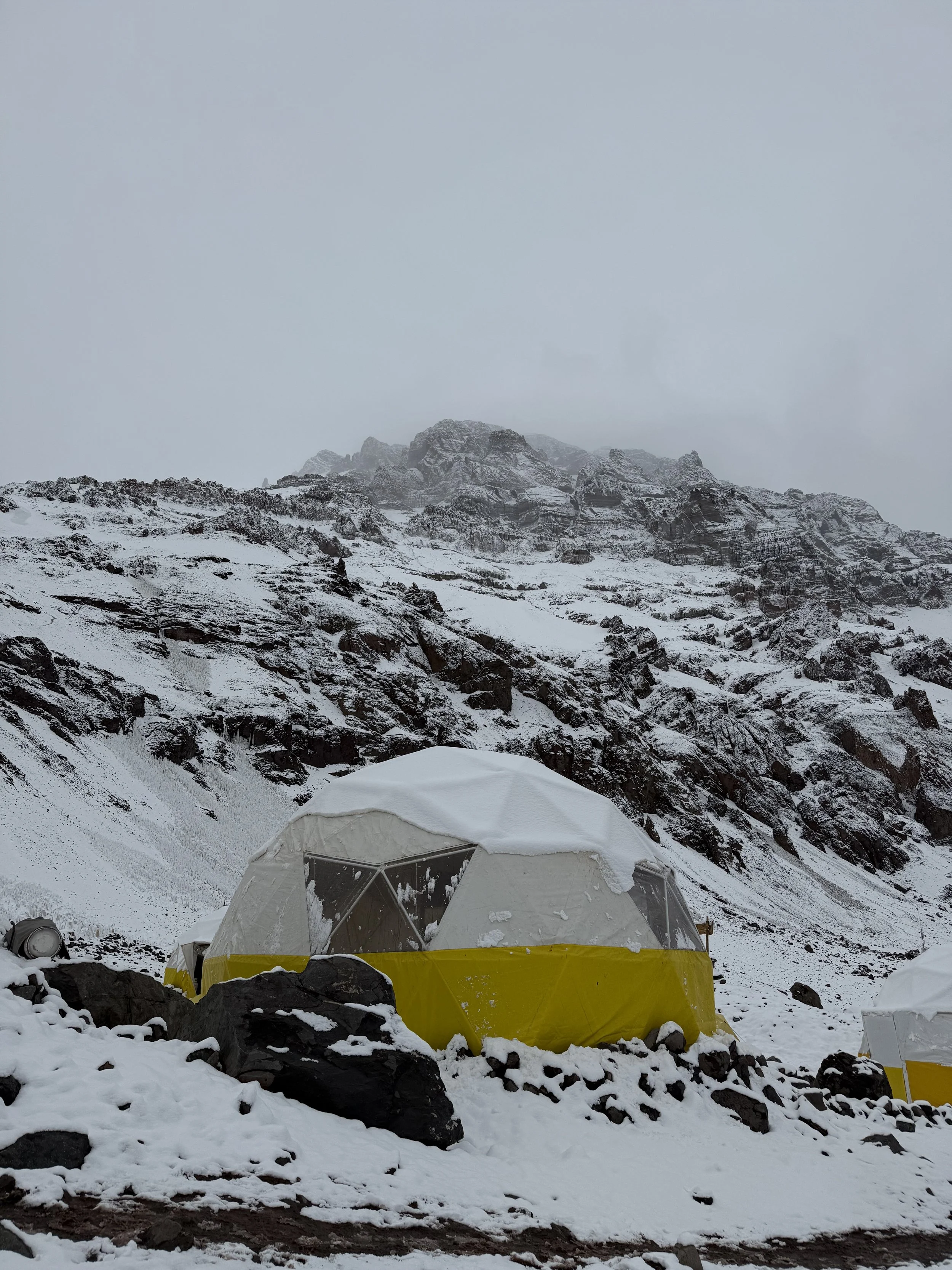 Yellow and white dome-shaped tent set up on snow-covered ground at the base of a steep, rocky, snow-covered mountain landscape.
