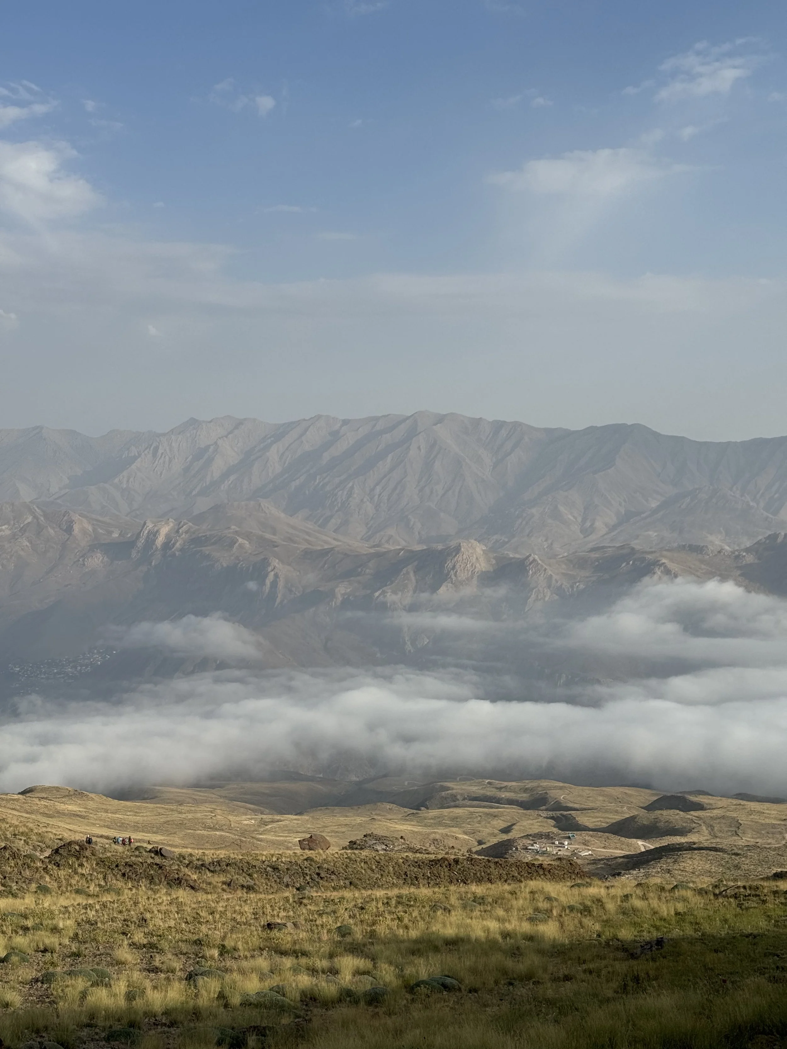 A landscape view of mountains with grassy plains in the foreground, cloud cover near the base of the mountains, and a partly cloudy sky.