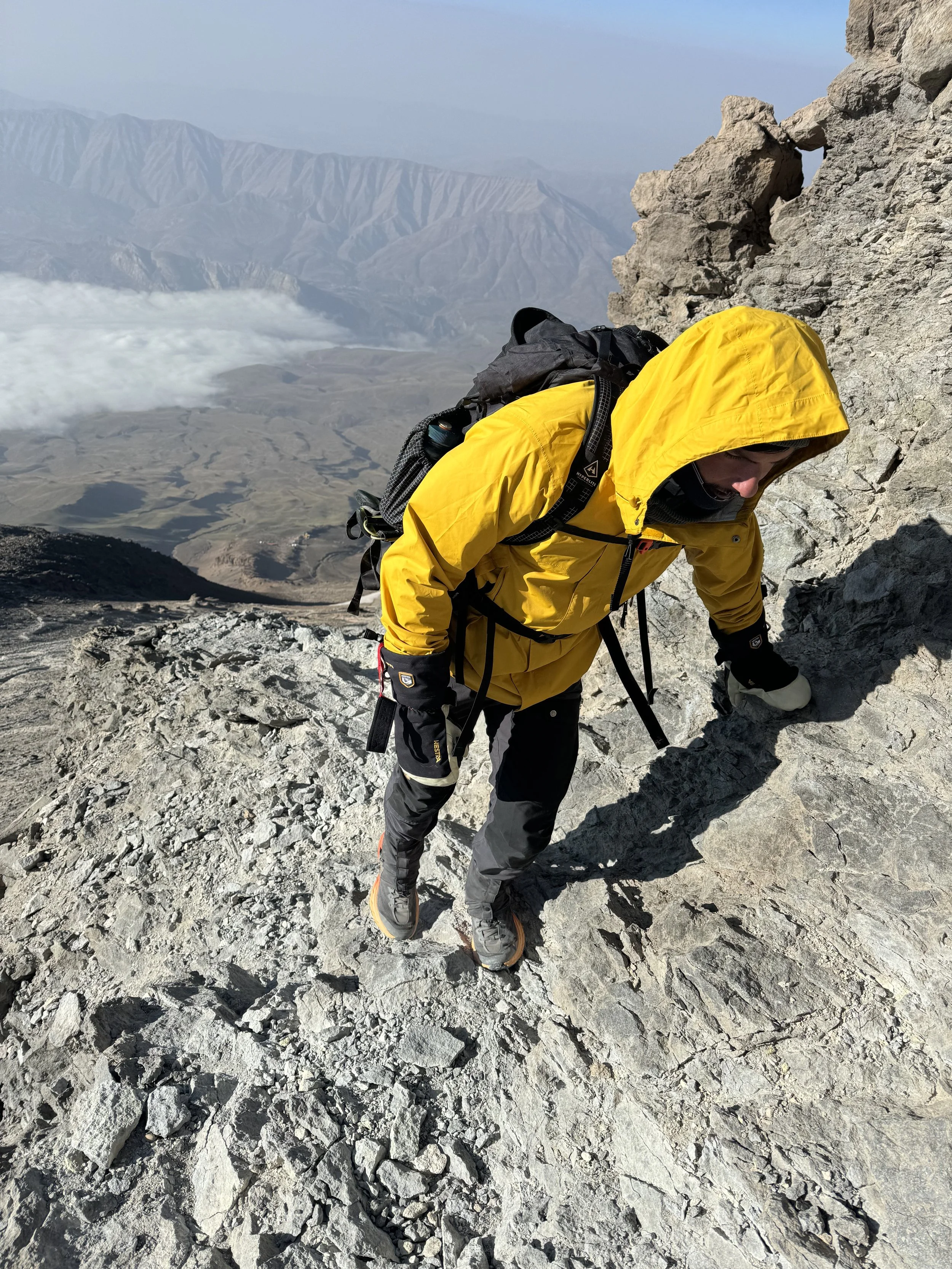 A man climbing a steep rocky mountain with a yellow rain jacket, black pants, and hiking boots, carrying a backpack. The mountain landscape and distant volcano are visible in the background.