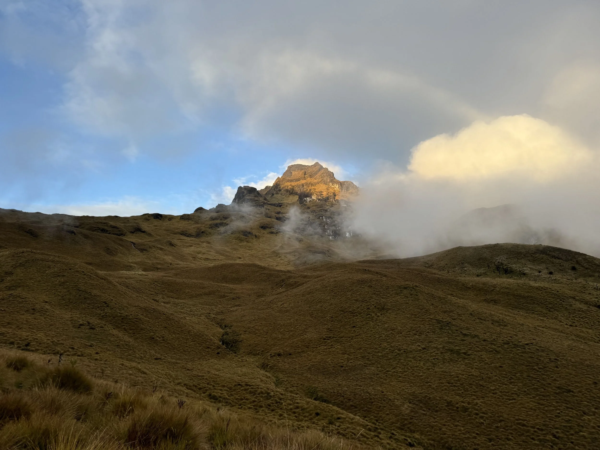 Rolling hills with grassy terrain leading to a mountain peak partly shrouded in clouds and mist, with a partly cloudy sky overhead.