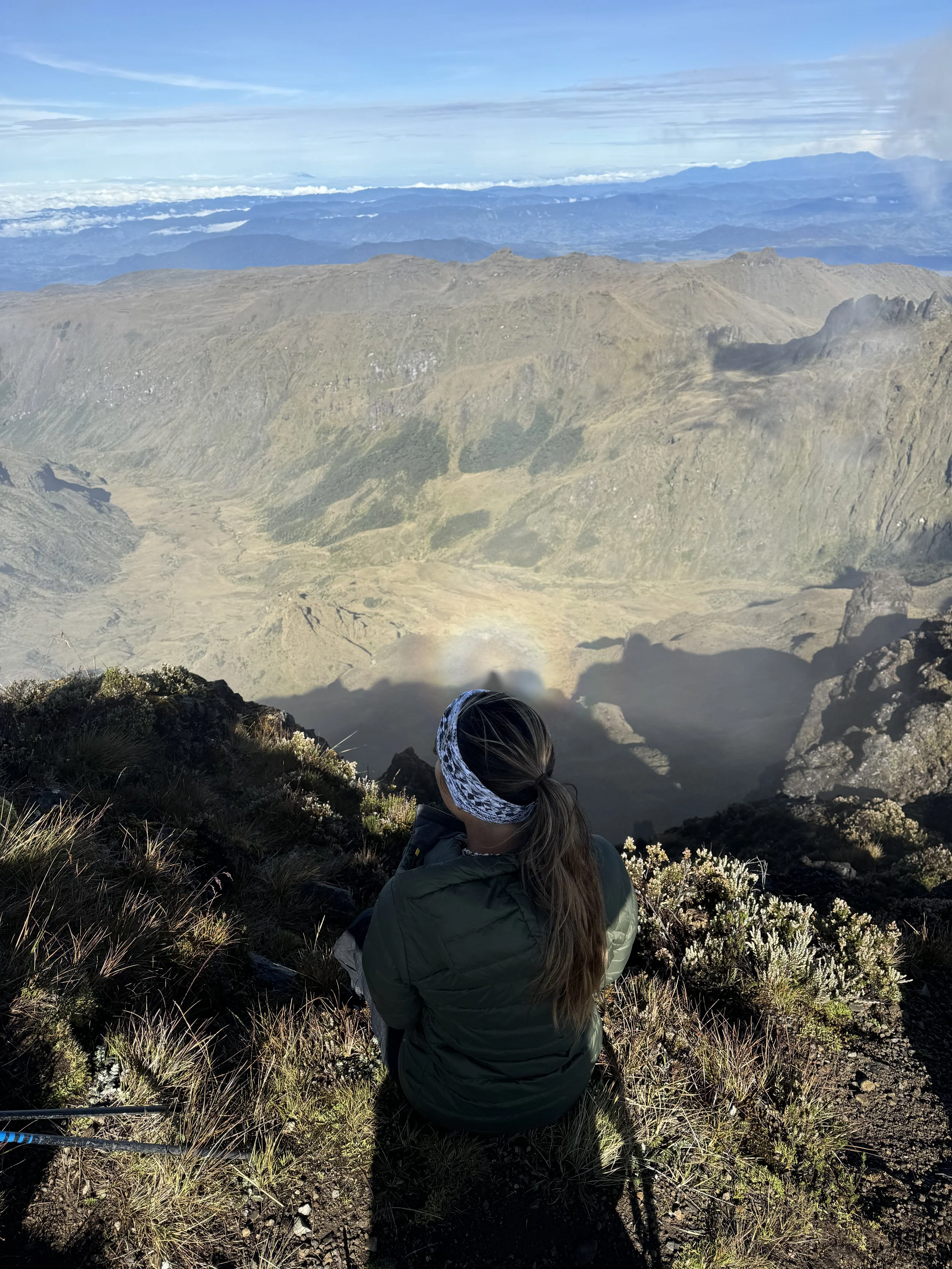 A woman sitting on a mountain trail overlooking a volcanic crater with a rainbow inside it, mountain ranges in the background, wearing a headband and a green jacket.