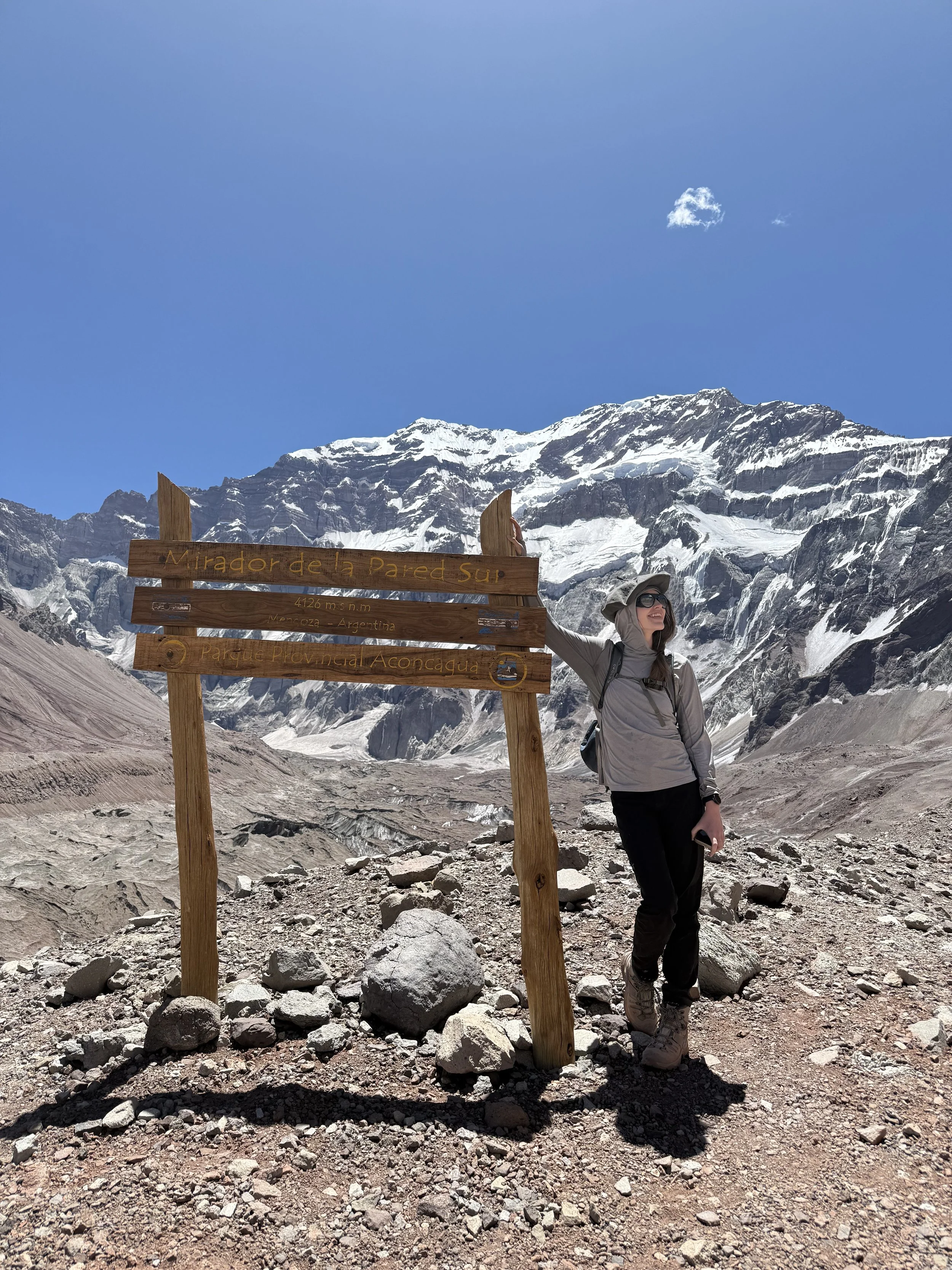 A woman with outdoor hiking gear standing next to a wooden sign in a mountainous, snowy landscape at high altitude. The sign reads "Mirador de la Pared Sur" with an elevation of 4,126 meters. Snow-capped mountains are visible in the background under