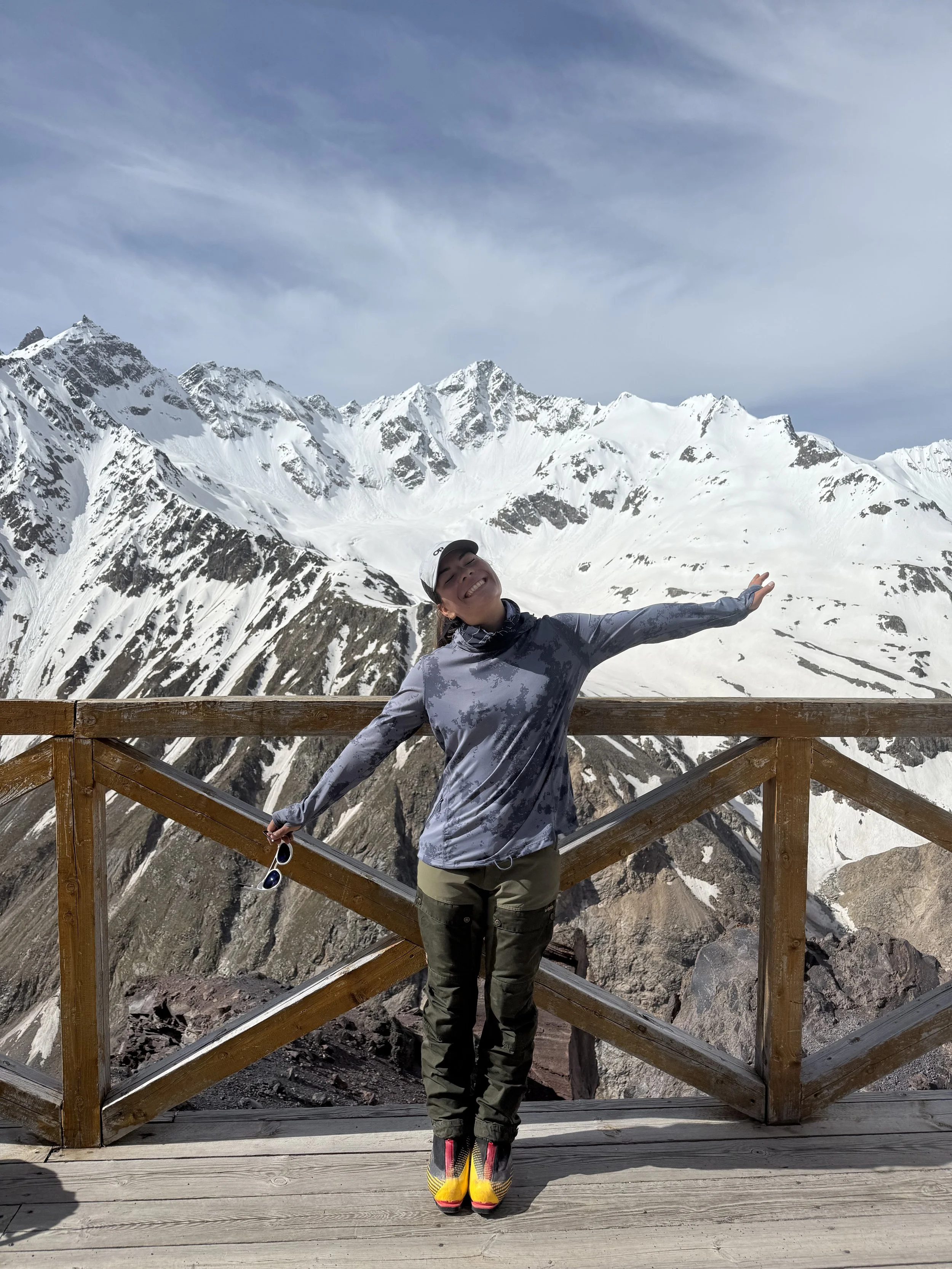 A woman standing on a wooden platform with a mountainous snowy landscape in the background, arms outstretched, smiling, holding sunglasses, wearing outdoor gear and colorful shoes.