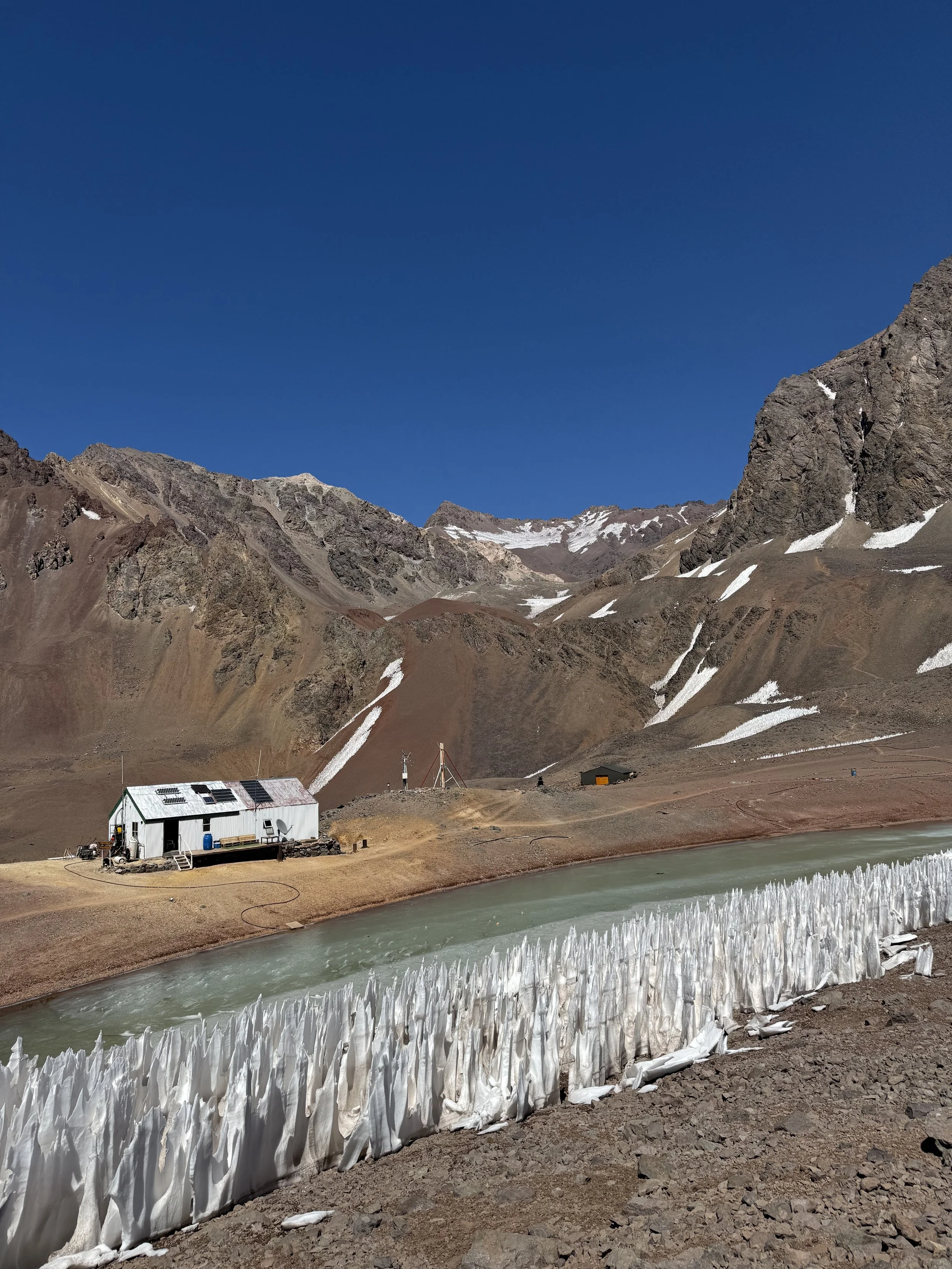 A small white building near a frozen river with high ice formations, set against rugged mountain terrain with patches of snow under a clear blue sky.