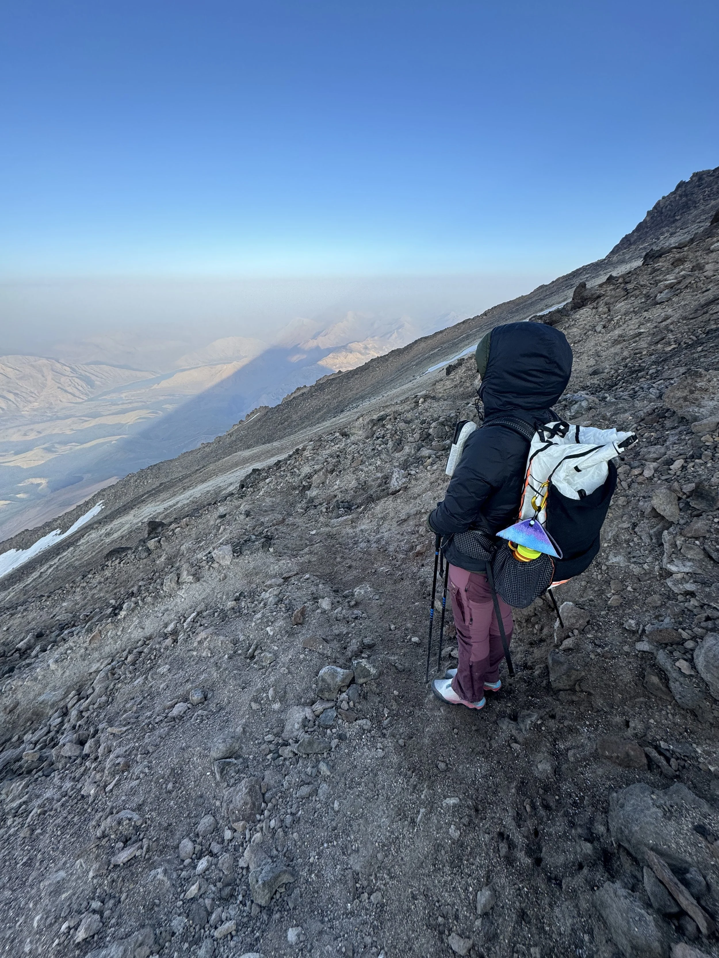 A person dressed in hiking gear, including a hooded jacket and hiking boots, stands on a rugged mountain trail with snow patches, overlooking a distant mountain landscape under a clear blue sky.
