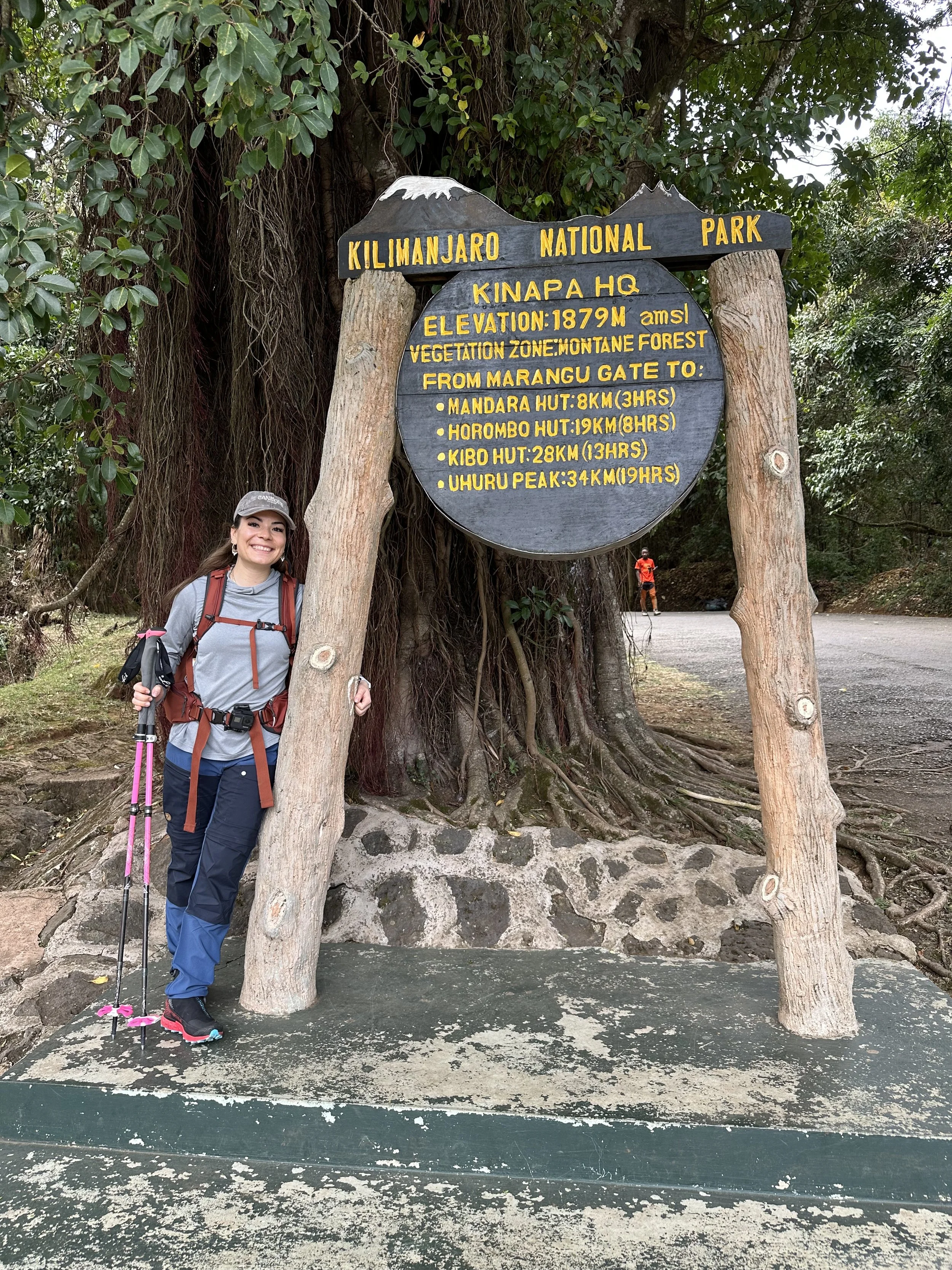 A woman in outdoor hiking gear and a hat standing next to a wooden signboard at Kilimanjaro National Park, Tanzania, indicating mountain information and hike distances. The sign is attached to two wooden posts in front of a large tree.