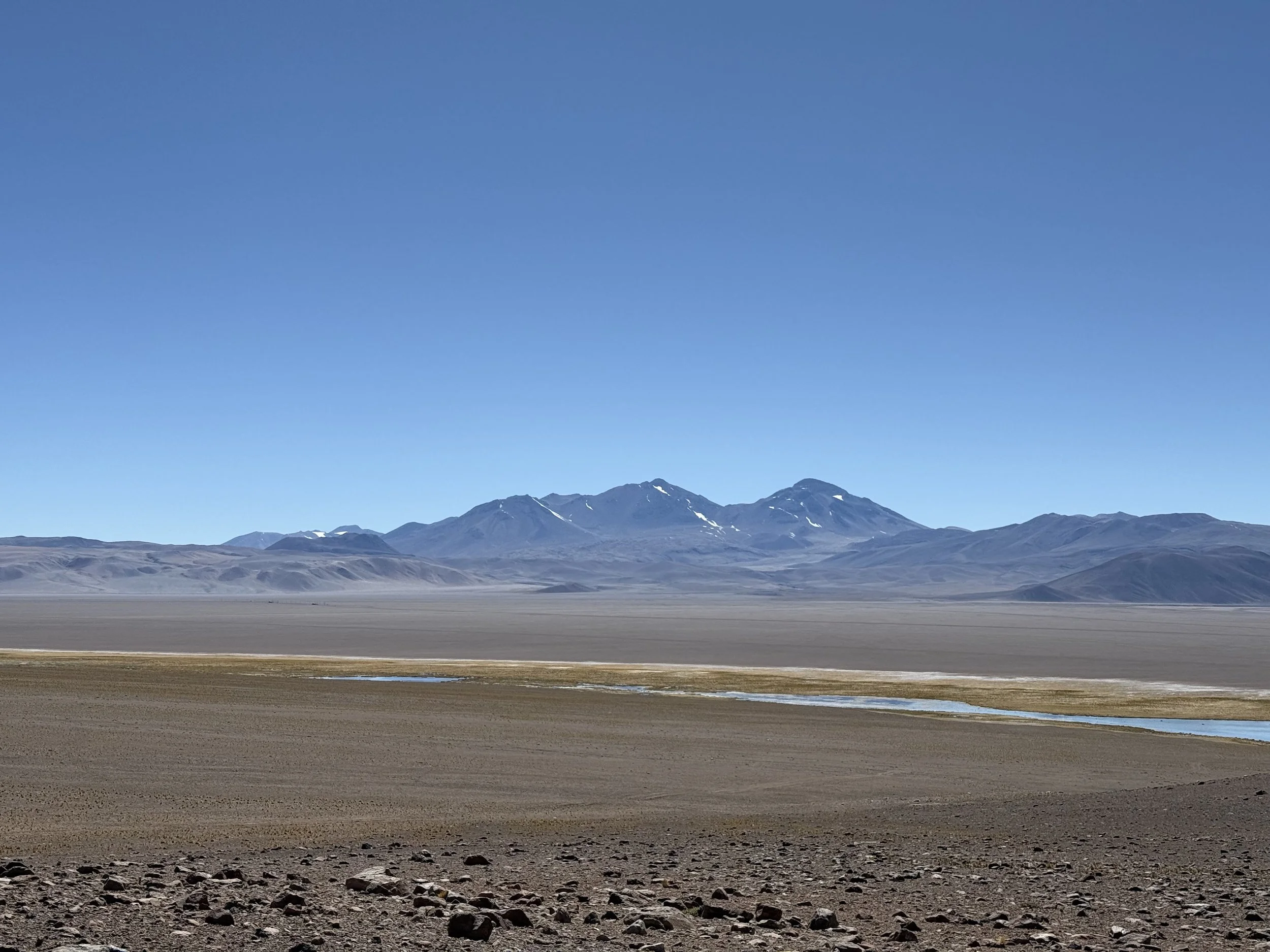 A vast desert landscape with brown rocky ground, a flat plain with some water bodies, distant mountains with snow patches, and a clear blue sky.