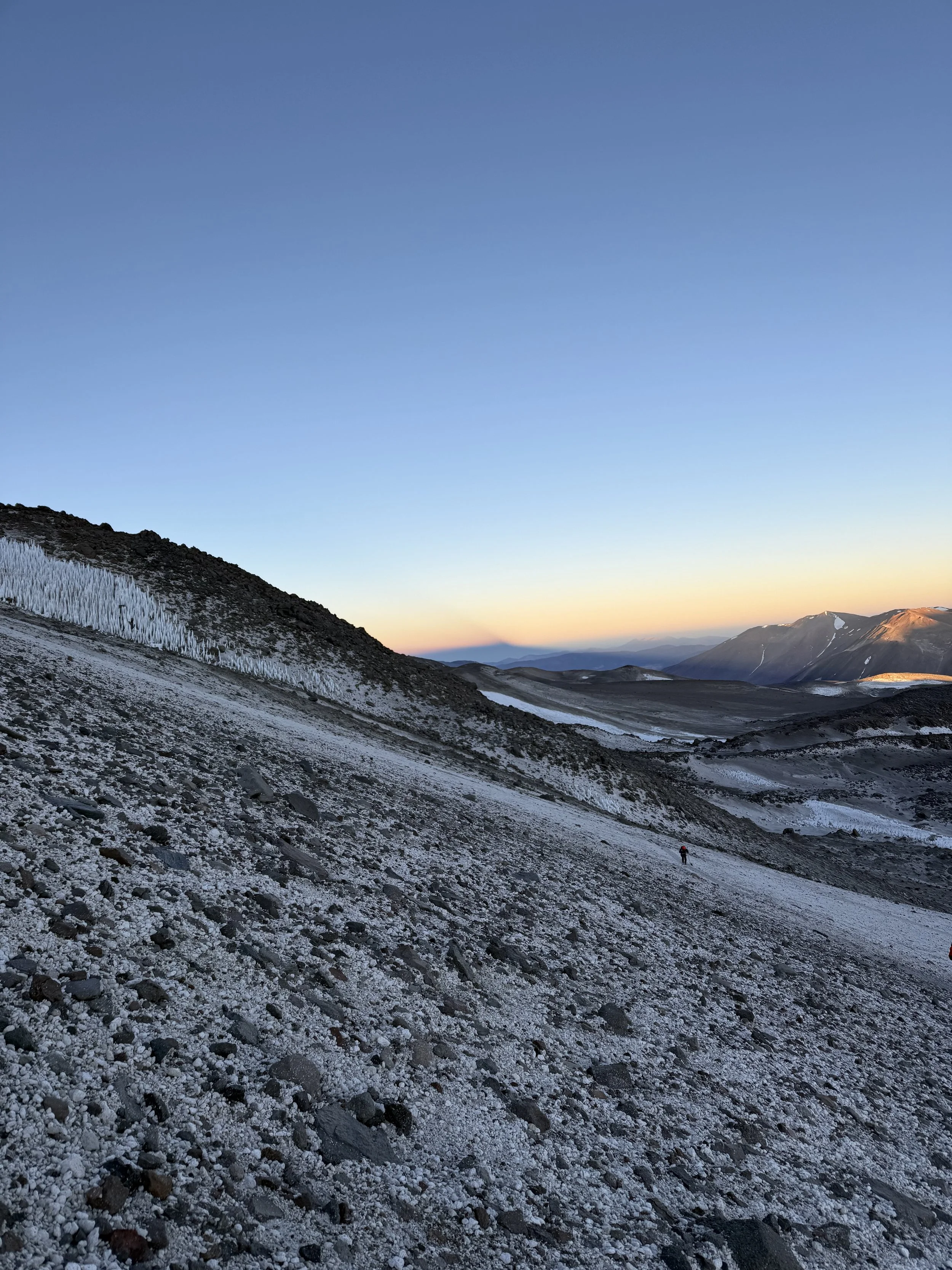 A mountain landscape with a clear sky, snow-covered terrain, and a lone hiker in the distance.