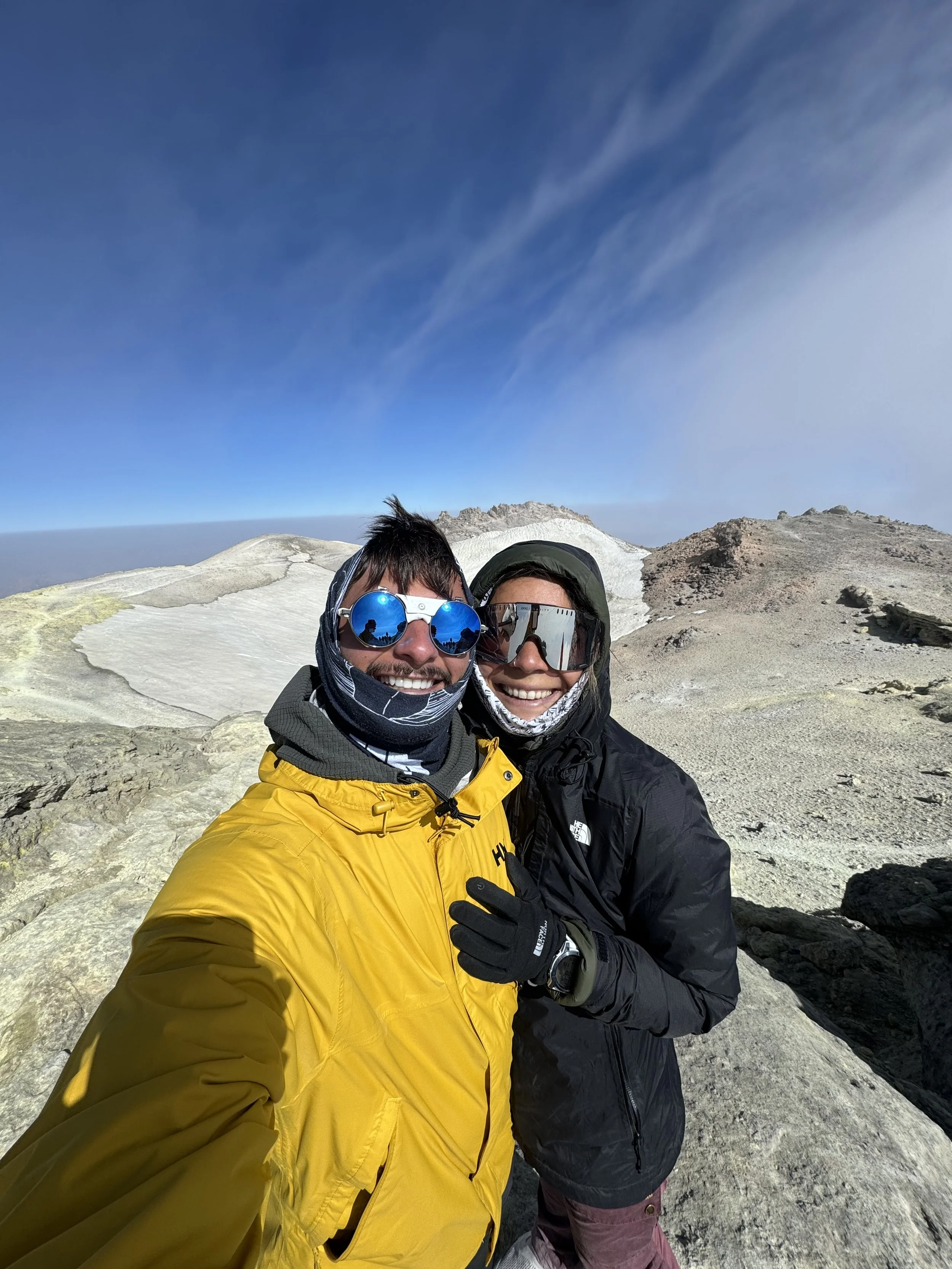 Two people wearing outdoor winter gear smiling on a mountain summit with volcanic rocks and a cloudy sky.