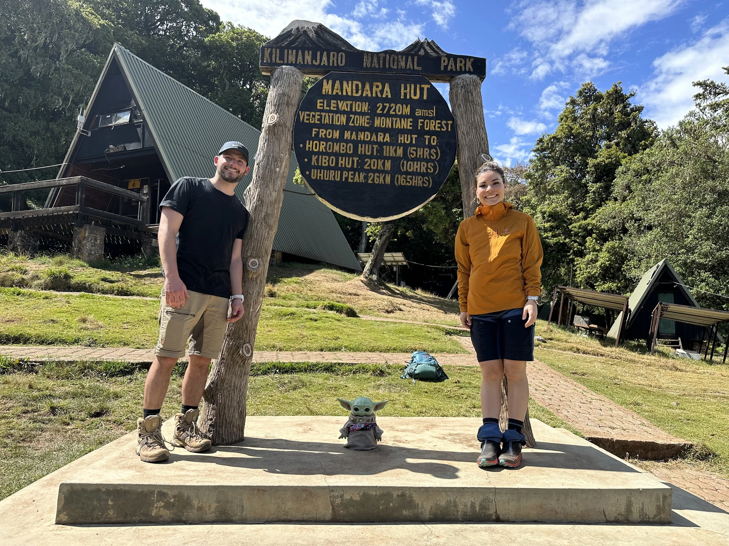 Two hikers, a man and a woman, stand beside a sign at Kilimanjaro National Park, indicating the Mandara Hut at 2720 meters elevation. The man is dressed in black t-shirt and beige shorts, while the woman wears an orange jacket and black shorts. A sma