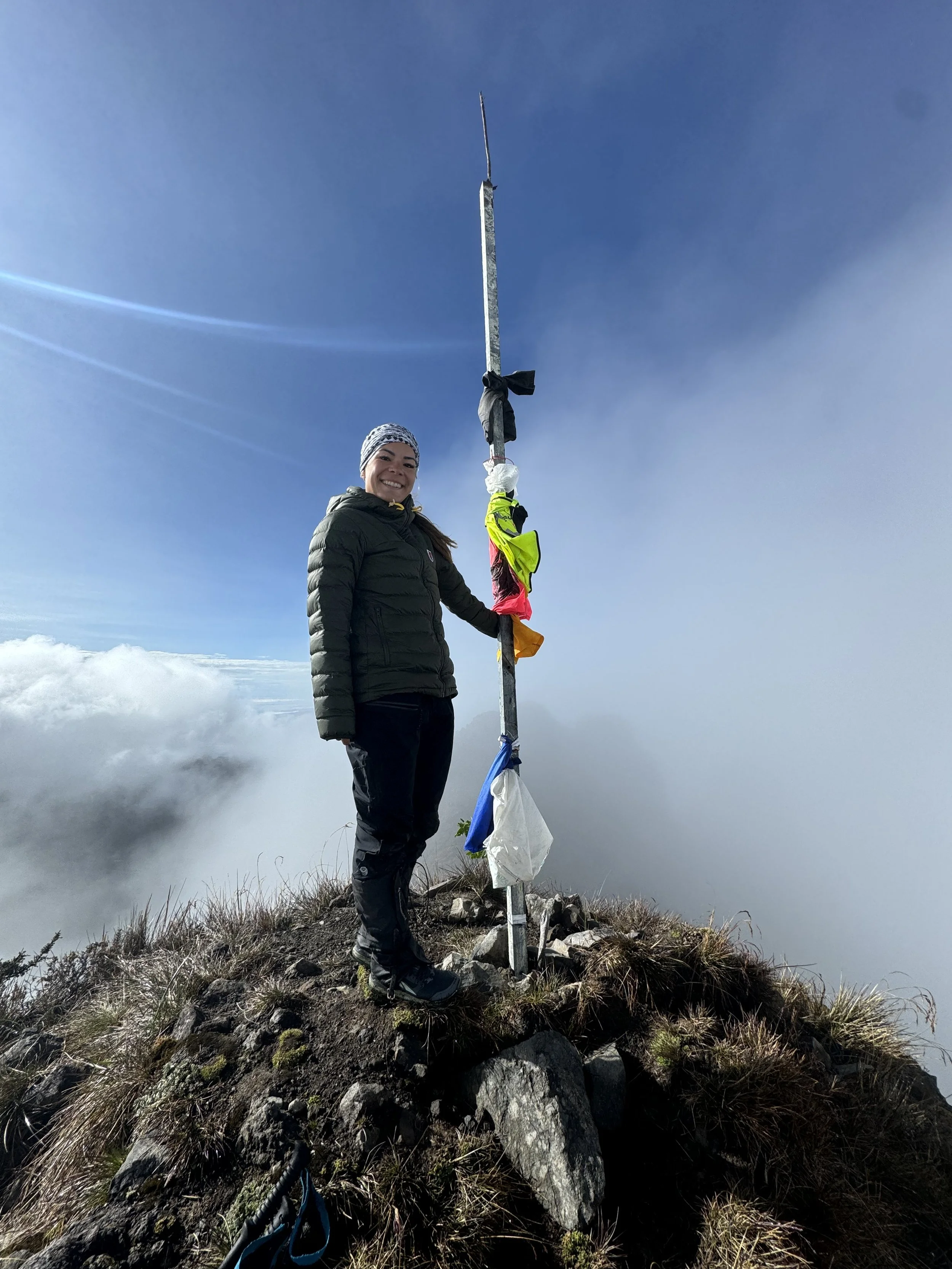 A woman stands on a mountain peak next to a tall summit marker with flags, clouds below, and a clear sky above.