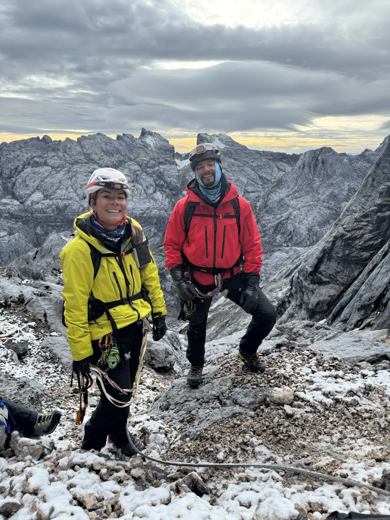 Two climbers in colorful jackets and helmets standing on rocky mountain terrain, with a rugged mountain range and cloudy sky in the background.
