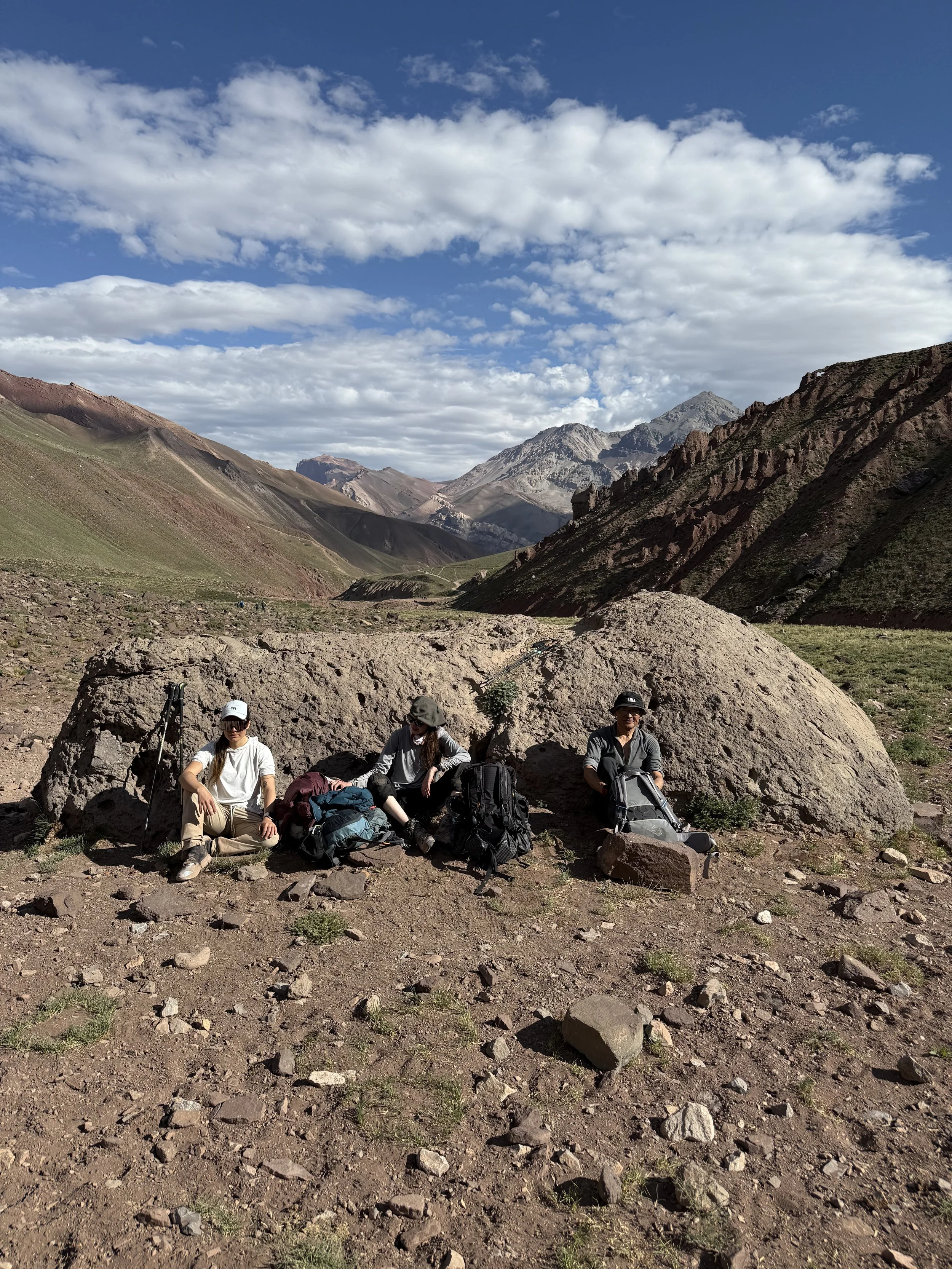 Three people taking a break while hiking in a mountainous area with rocky terrain, mountains in the background, and a partly cloudy sky.