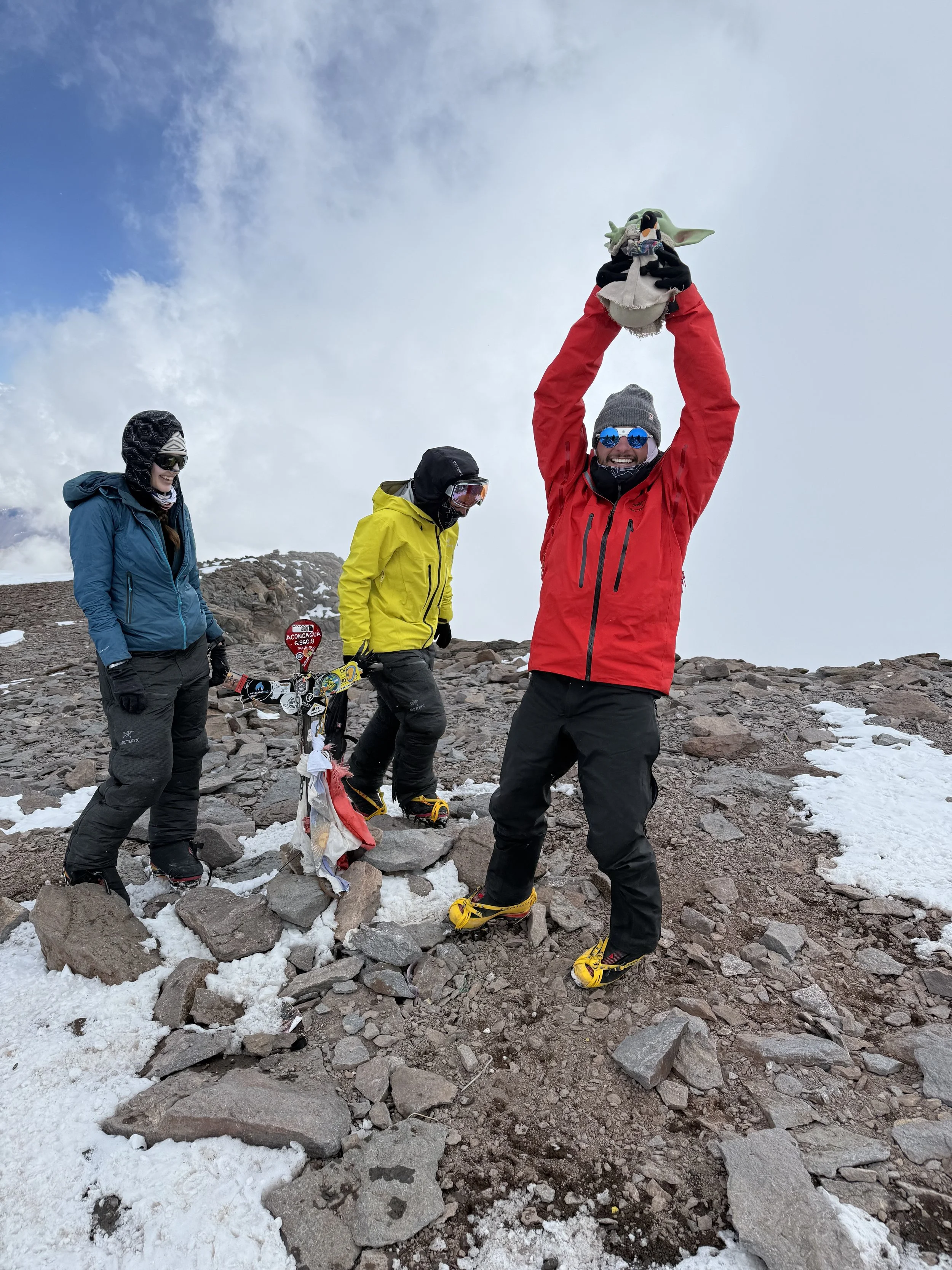 Three people in winter gear on a rocky, snowy mountain summit. One person wearing a red jacket is raising their arms, holding a green plush toy. The other two are standing nearby, one in a yellow jacket and the other in a blue jacket, with an America
