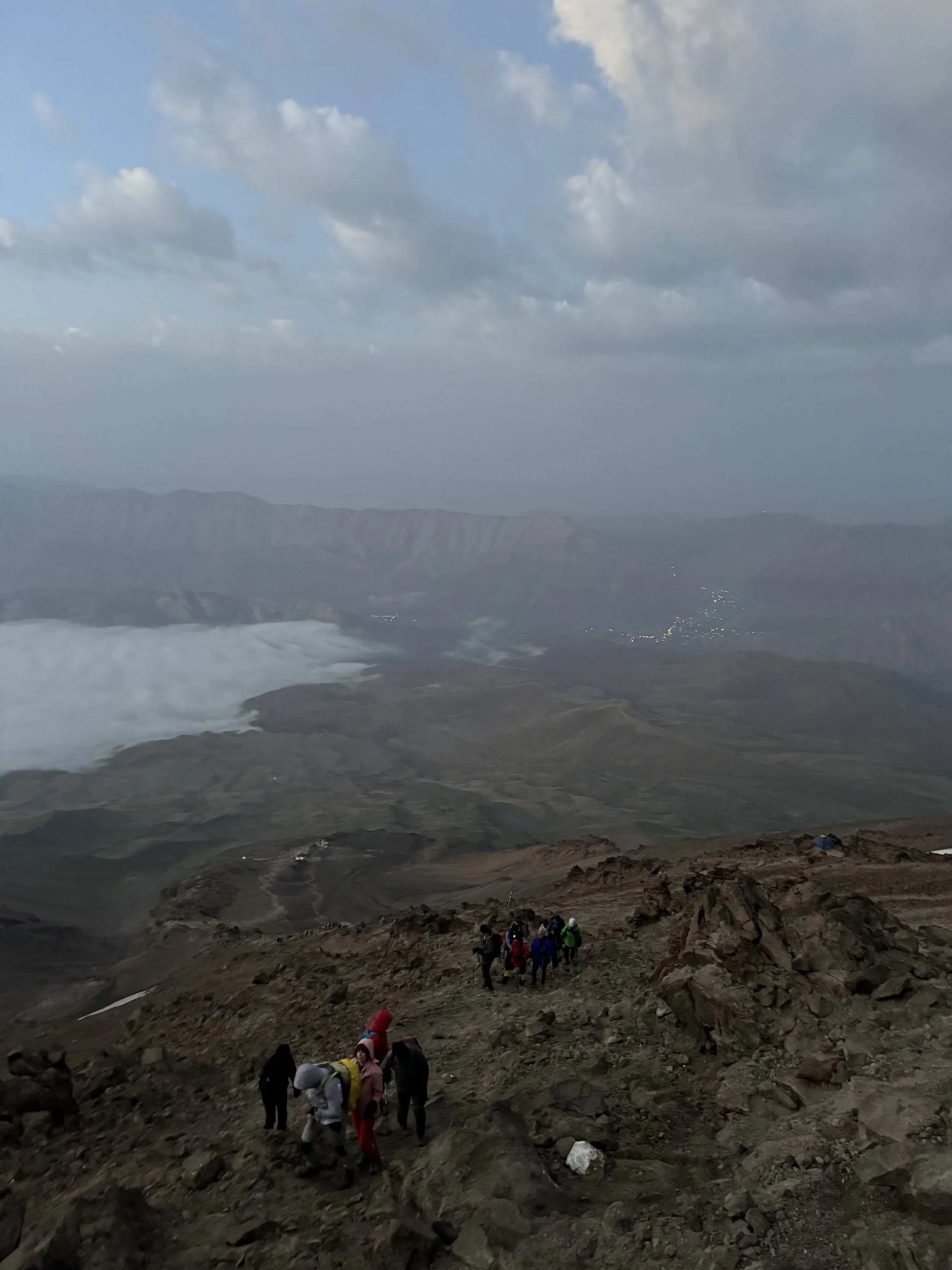 A group of hikers ascending a rocky mountain trail with a view of a valley, mountains, and clouds in the background.