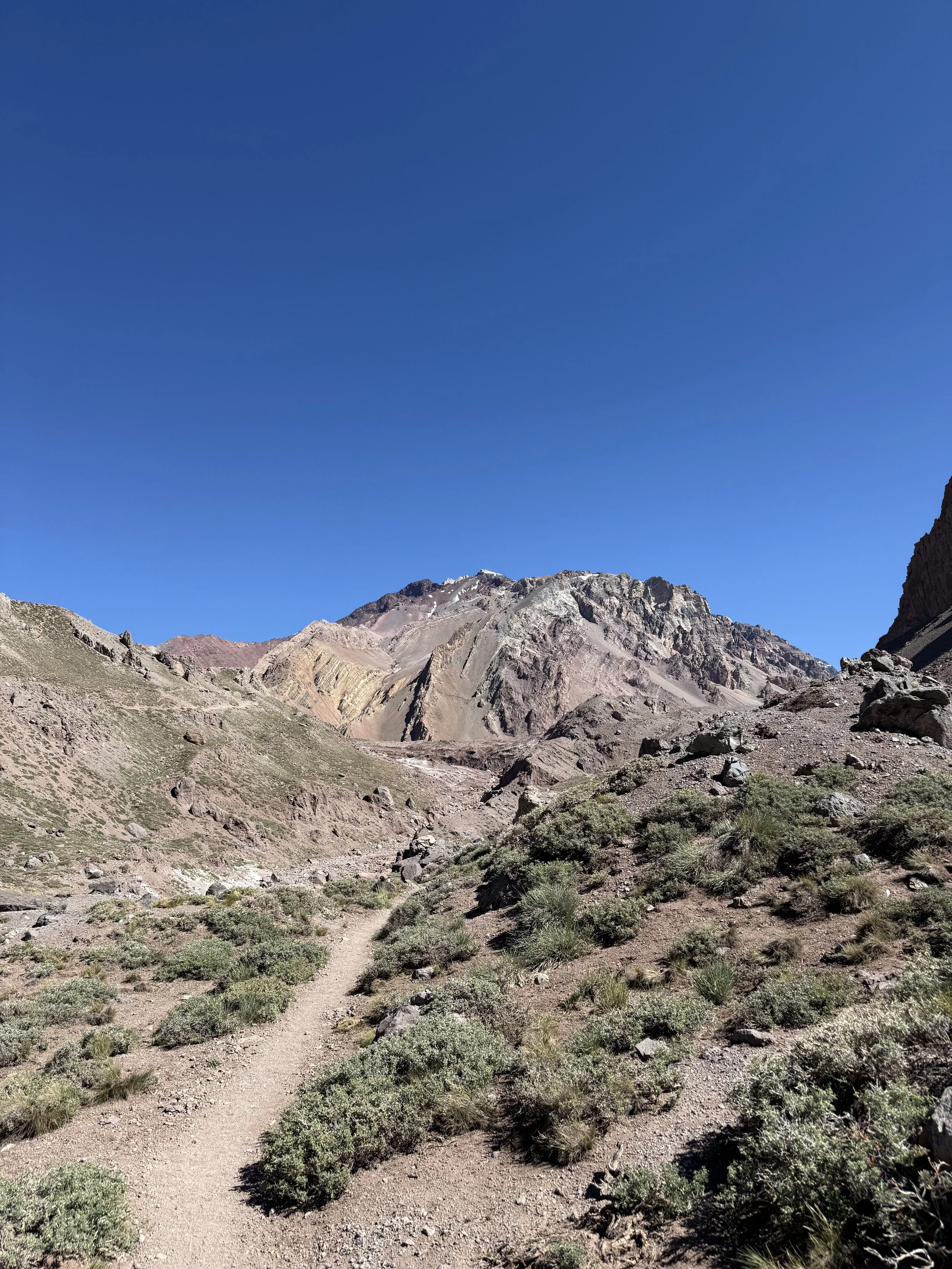 A rugged mountain landscape with a dirt trail leading towards rocky, multicolored mountains under a clear blue sky.