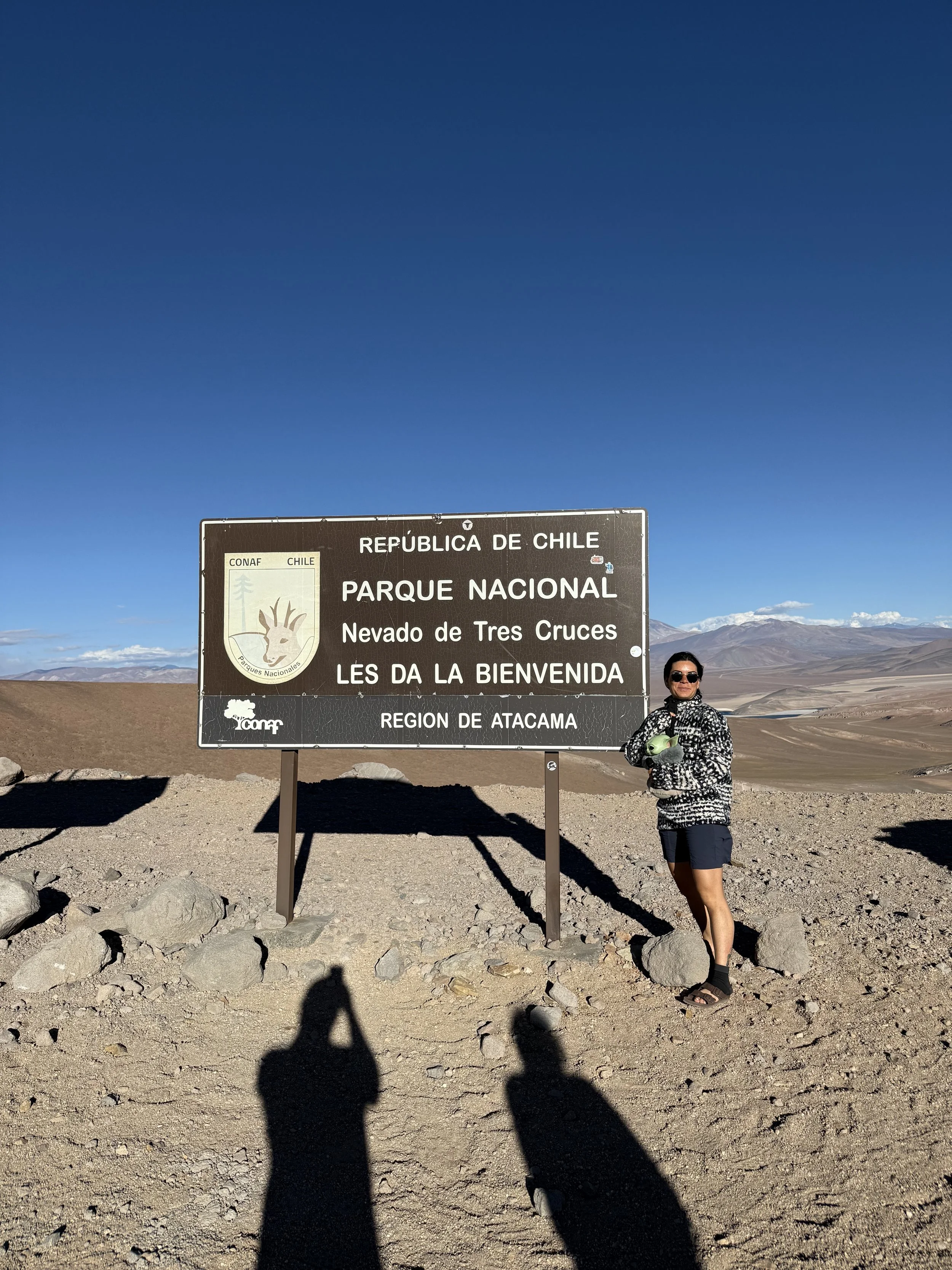 A woman dressed in black and white standing next to a large sign at the National Park Nevado de Tres Cruces in the Atacama region of Chile. The sign is brown with white lettering, indicating the location in Spanish and featuring a logo of the nationa