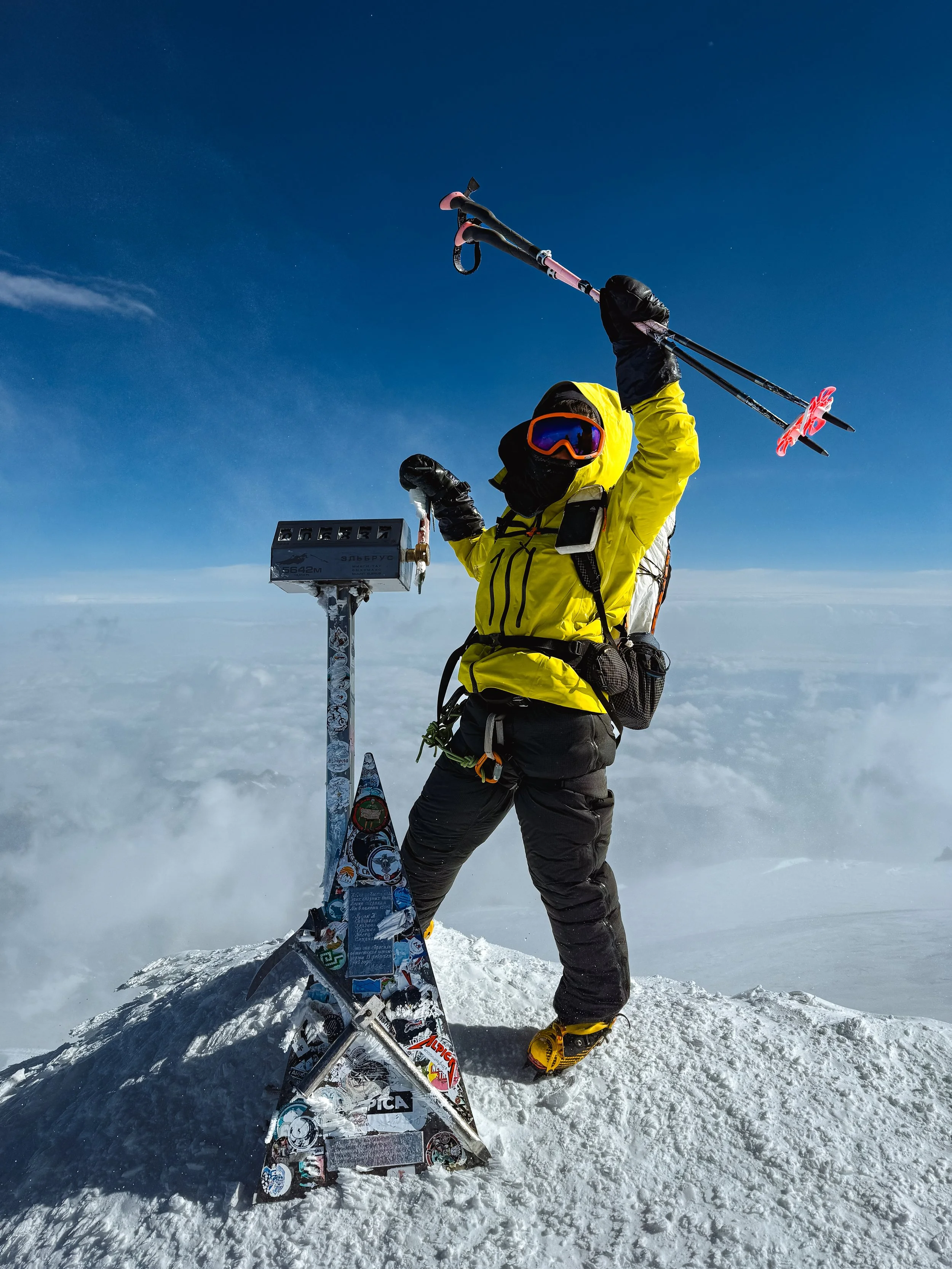 A mountaineer wearing a yellow jacket, black pants, and ski gear stands triumphantly on a snow-covered summit, holding ski poles raised in the air. The person is in front of a snow pole with a cloud-filled sky and blue sky in the background.