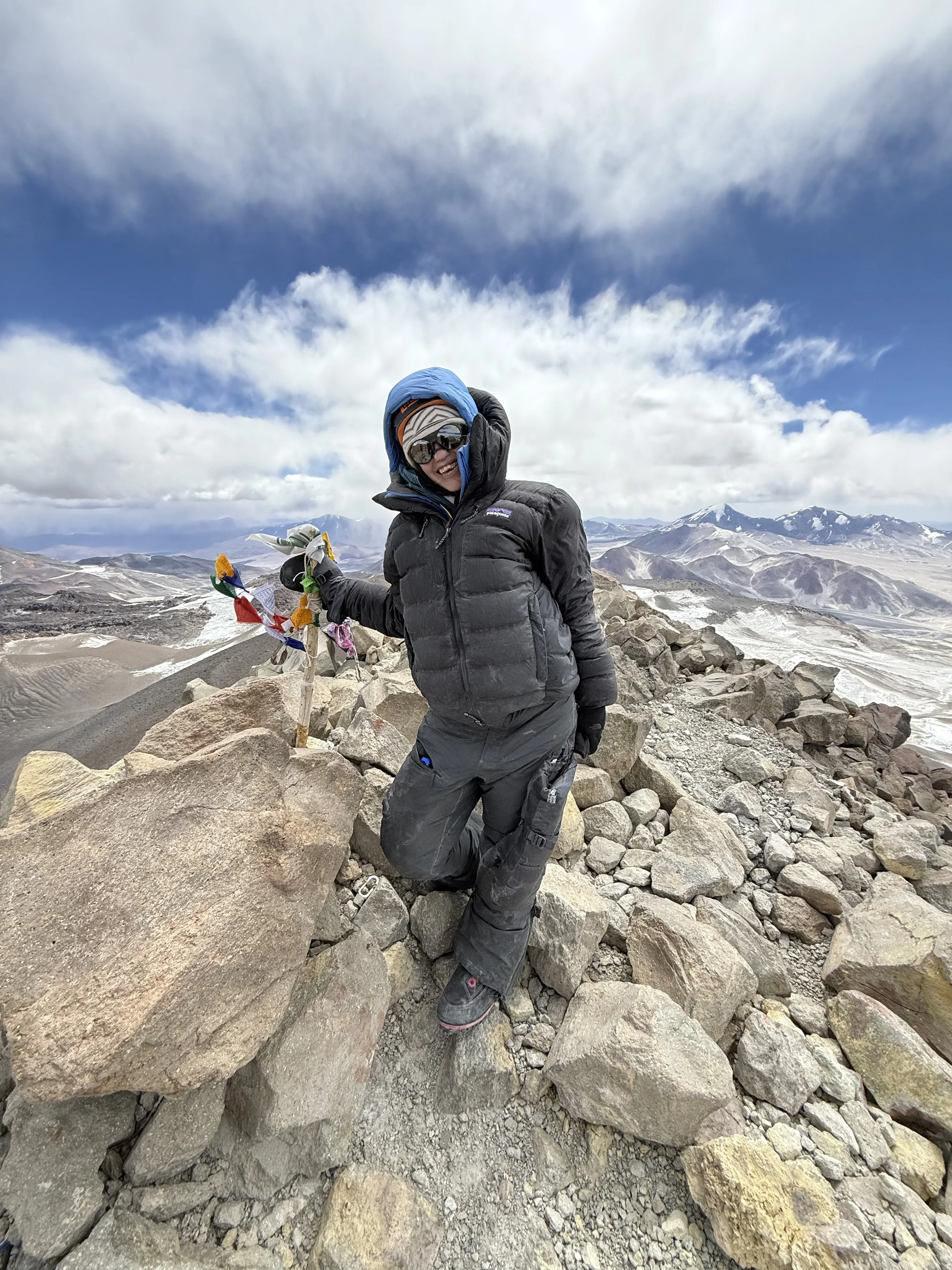 A person in outdoor winter gear, including a black jacket, pants, gloves, and a head covering, is kneeling on rocky terrain in a mountainous area. They are smiling and holding a small staff decorated with colorful cloths and ribbons. The background f