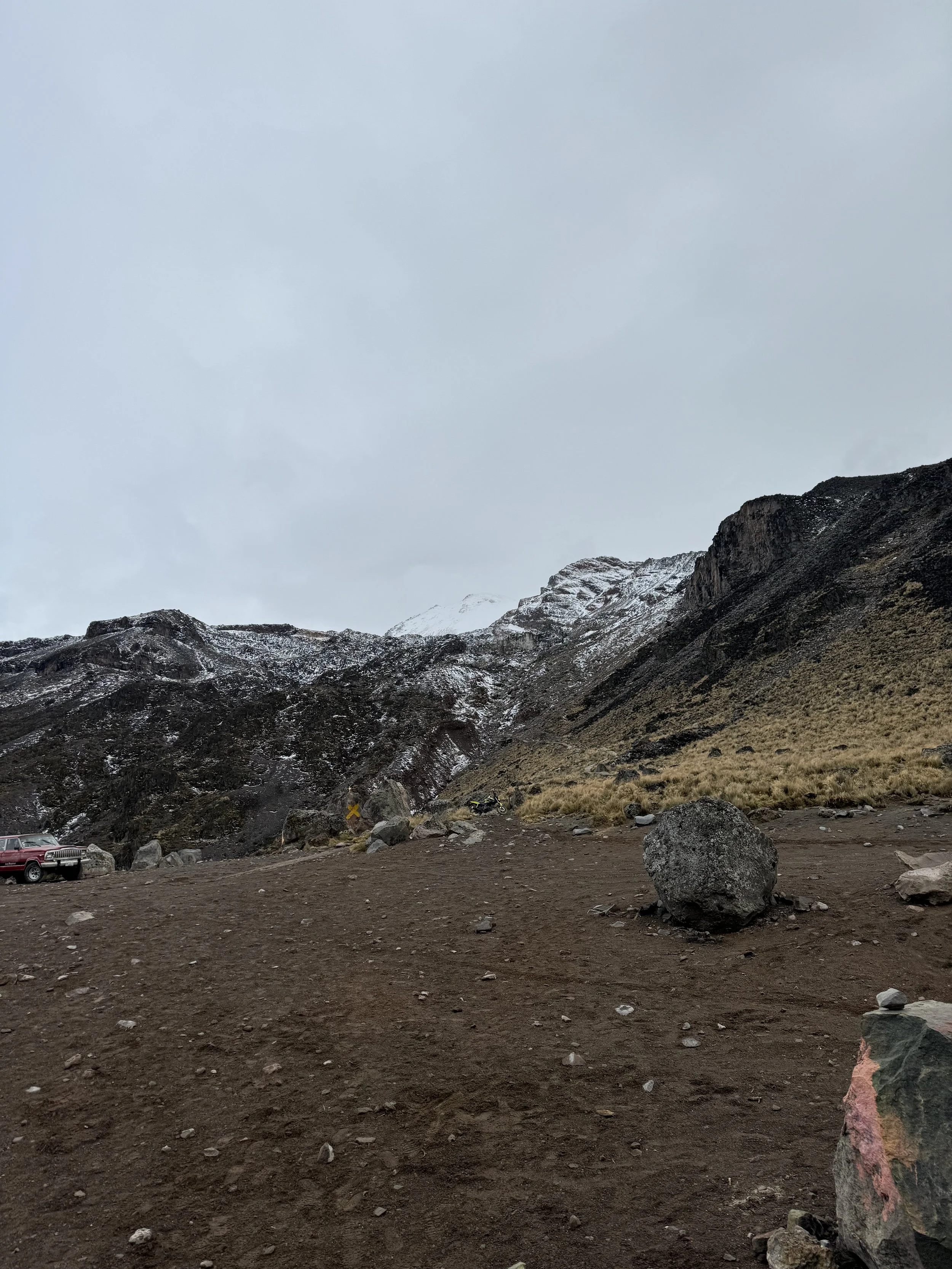 A rugged mountain landscape with brown dirt ground, large rocks, a yellow sign, a car, and snowy mountain peaks under a cloudy sky.