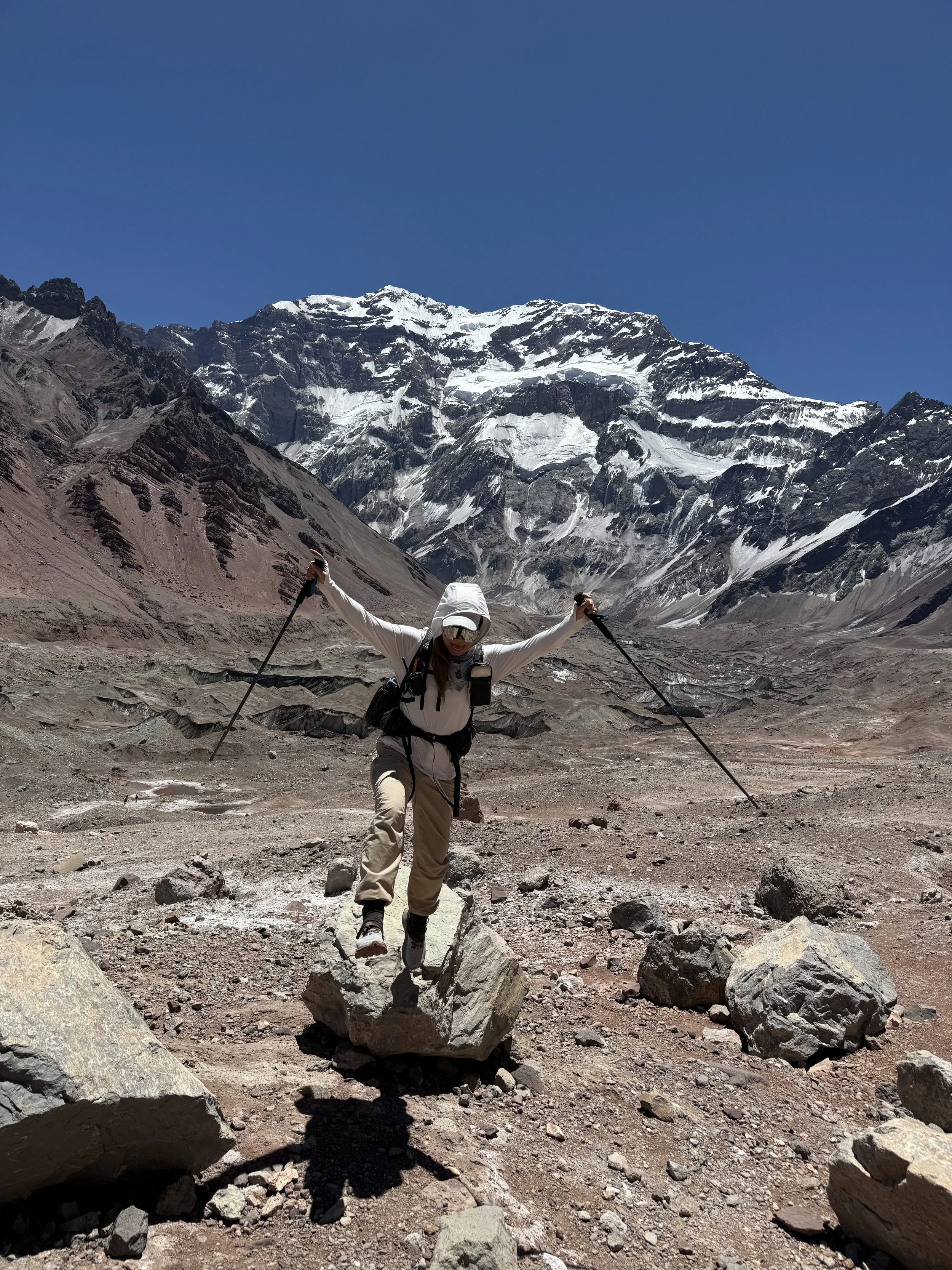 A person hiking in a barren rocky mountain landscape with snow-capped peaks in the background, wearing outdoor gear and holding trekking poles.