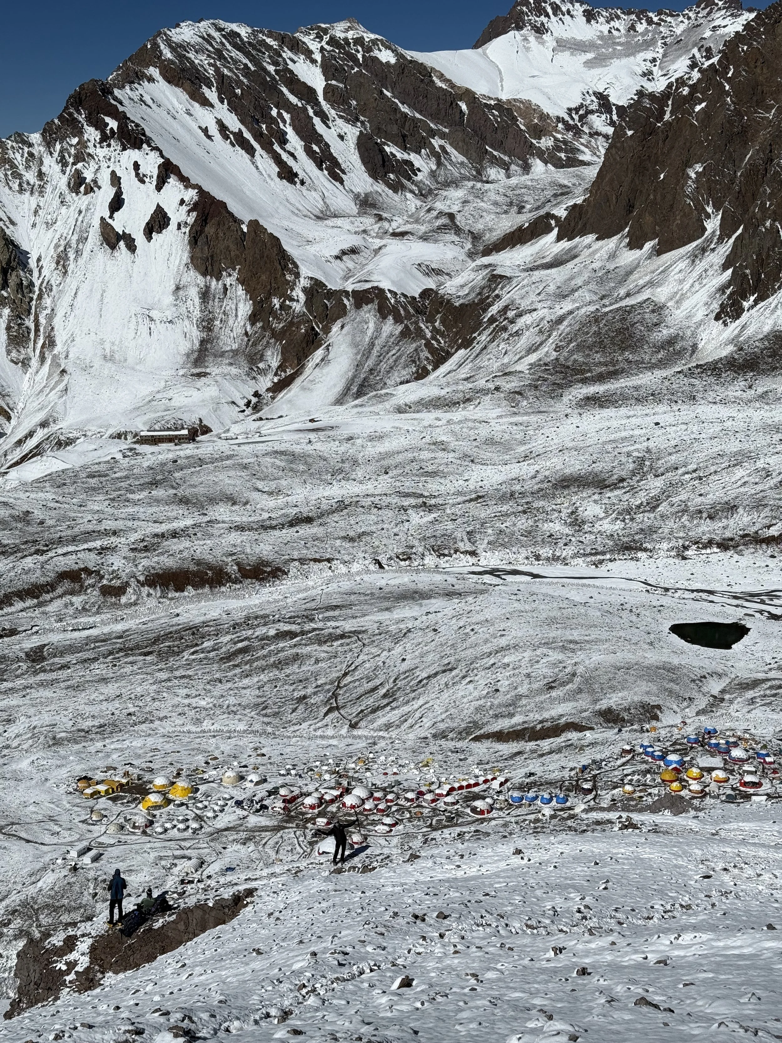 A snowy mountain landscape with a small encampment of colorful tents at the base of the mountain and a few people standing nearby.