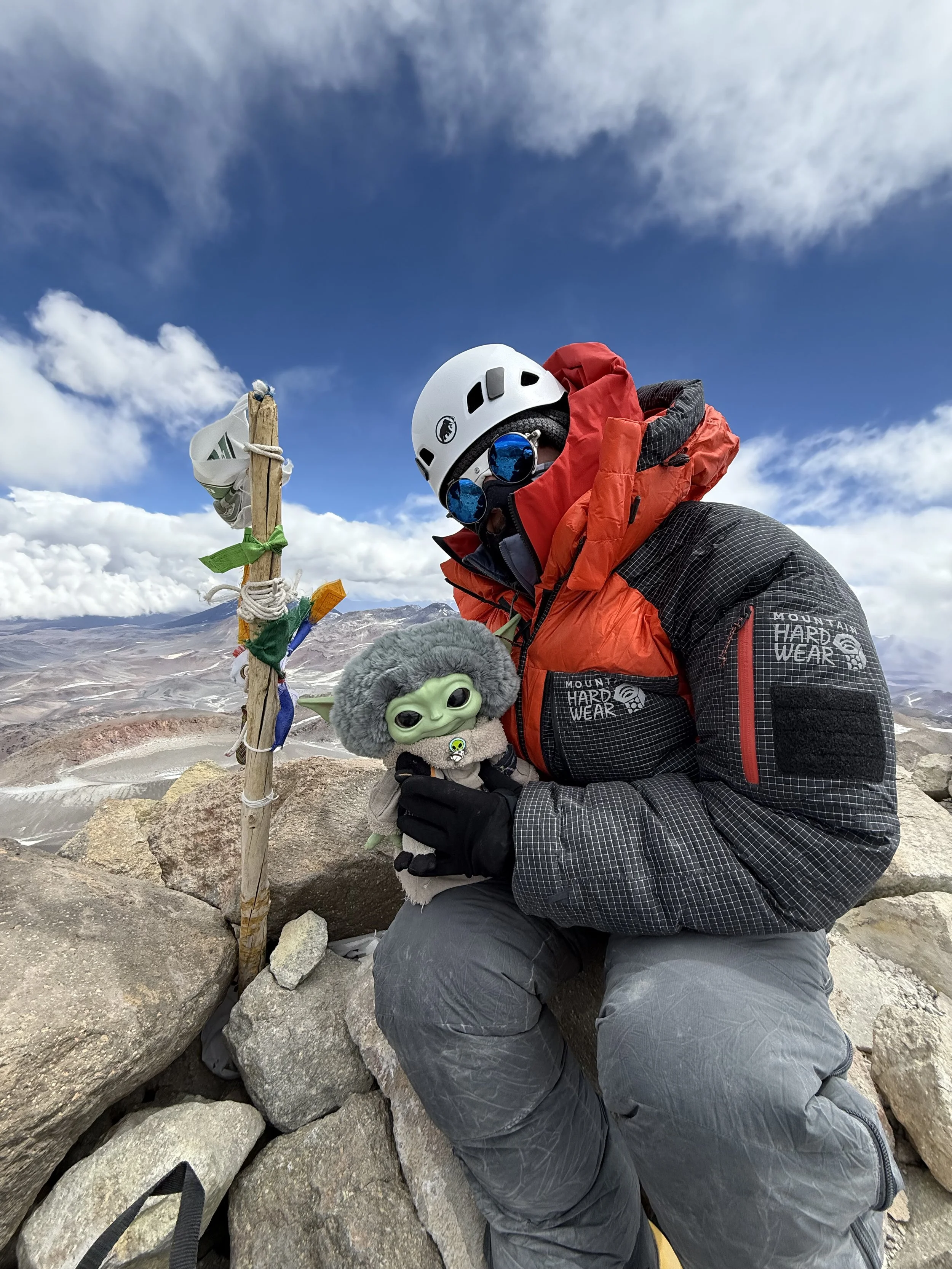 A hiker dressed in a red and black mountain jacket, gray pants, white helmet, blue sunglasses, and gloves, holding a plush toy of Baby Yoda with a gray hat and black clothing, kneeling on rocks in a mountainous landscape with a flagpole wrapped with