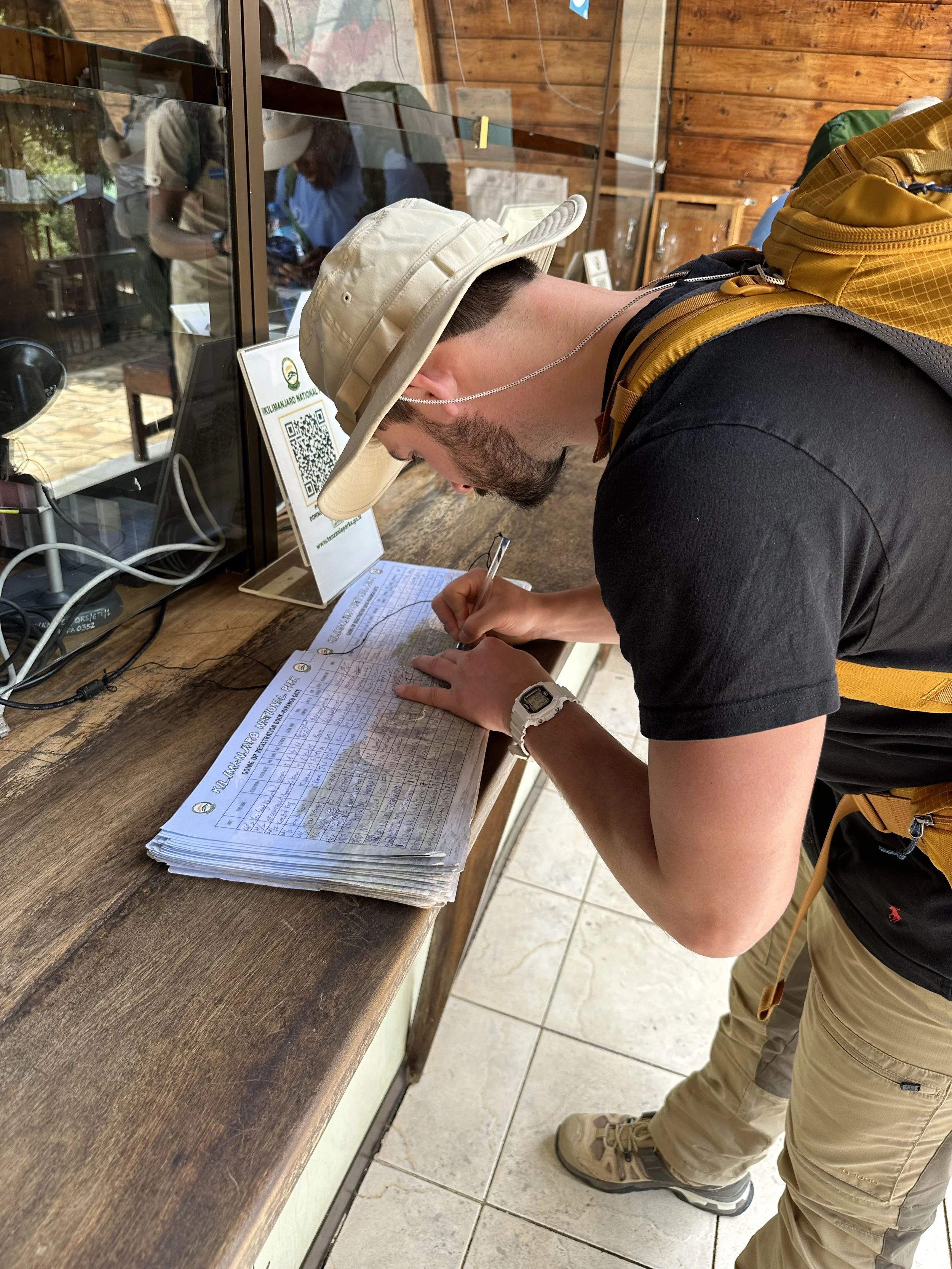 A man wearing a beige hat, black t-shirt, tan cargo pants, and a yellow backpack is signing a document on a wooden table. He is wearing a watch and is leaning over the table, filling out paperwork.
