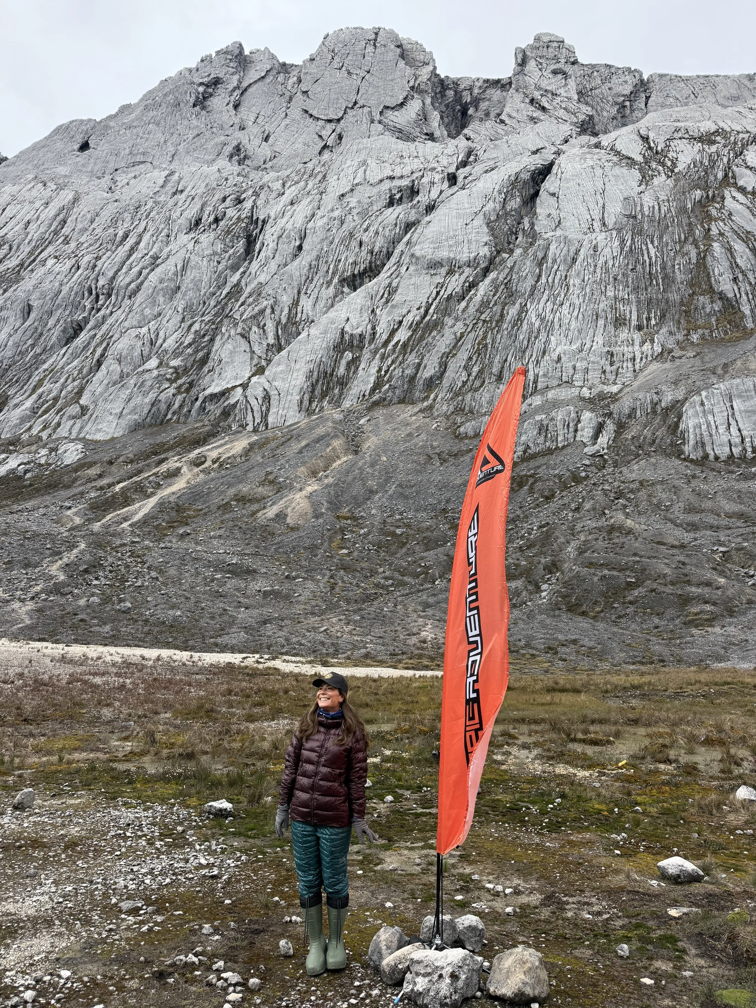 A woman in outdoor gear standing on rocky terrain near a tall orange flag with black text, with a large gray mountain in the background.