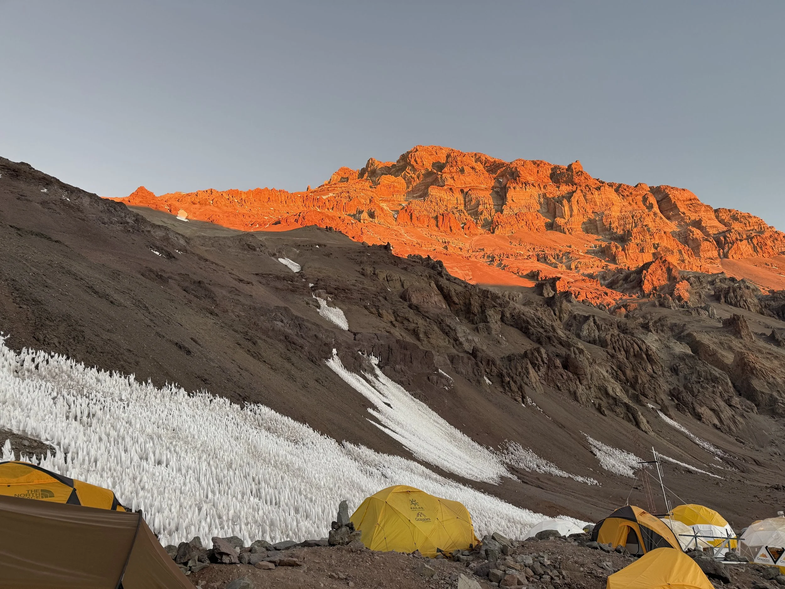 Yellow tents set up on rocky ground at the base of a mountain with orange-lit summit, patches of white snow, and a clear sky.