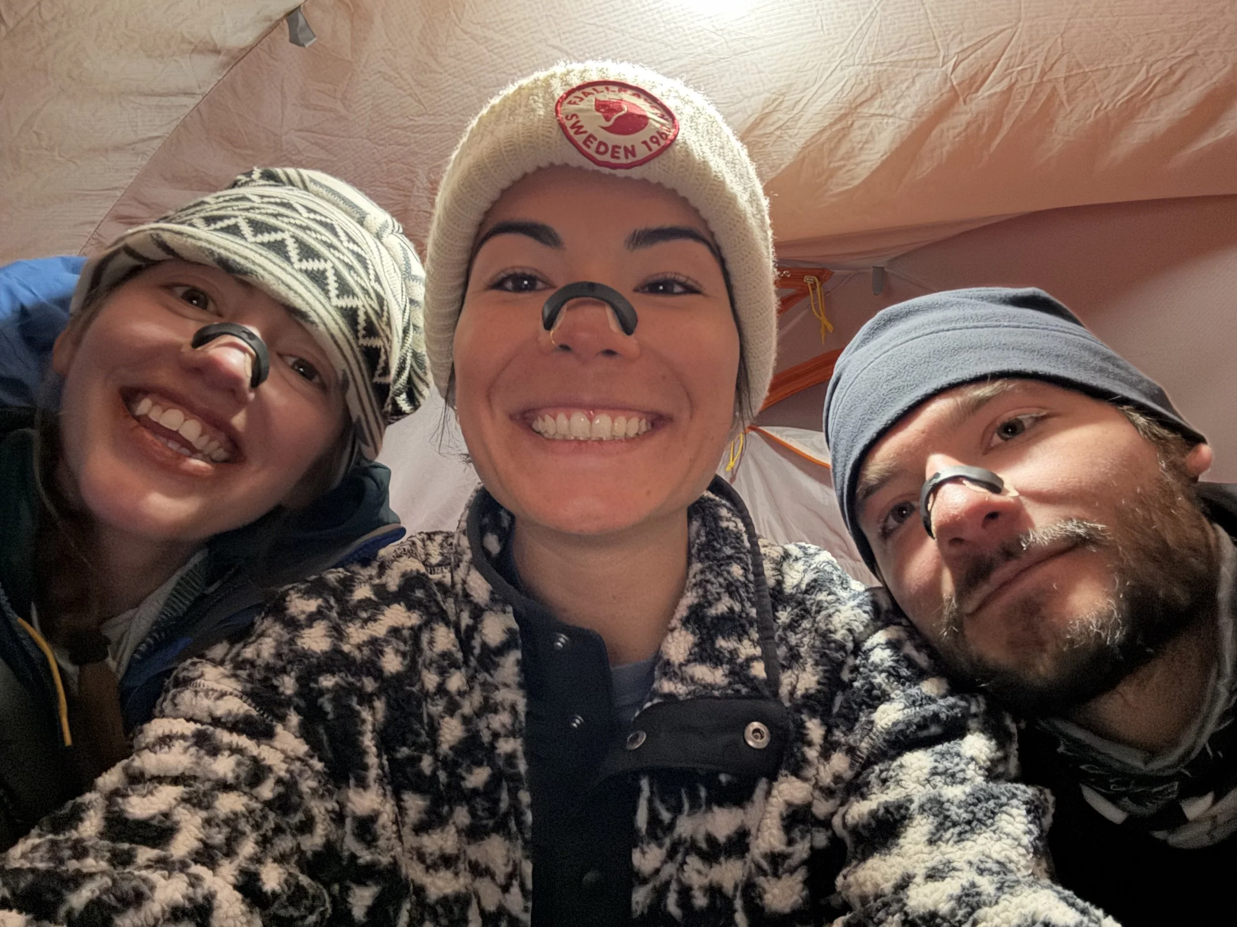 Three smiling friends inside a tent, wearing warm clothing, beanies, and headbands, with one person in the middle wearing a beanie with a red logo.