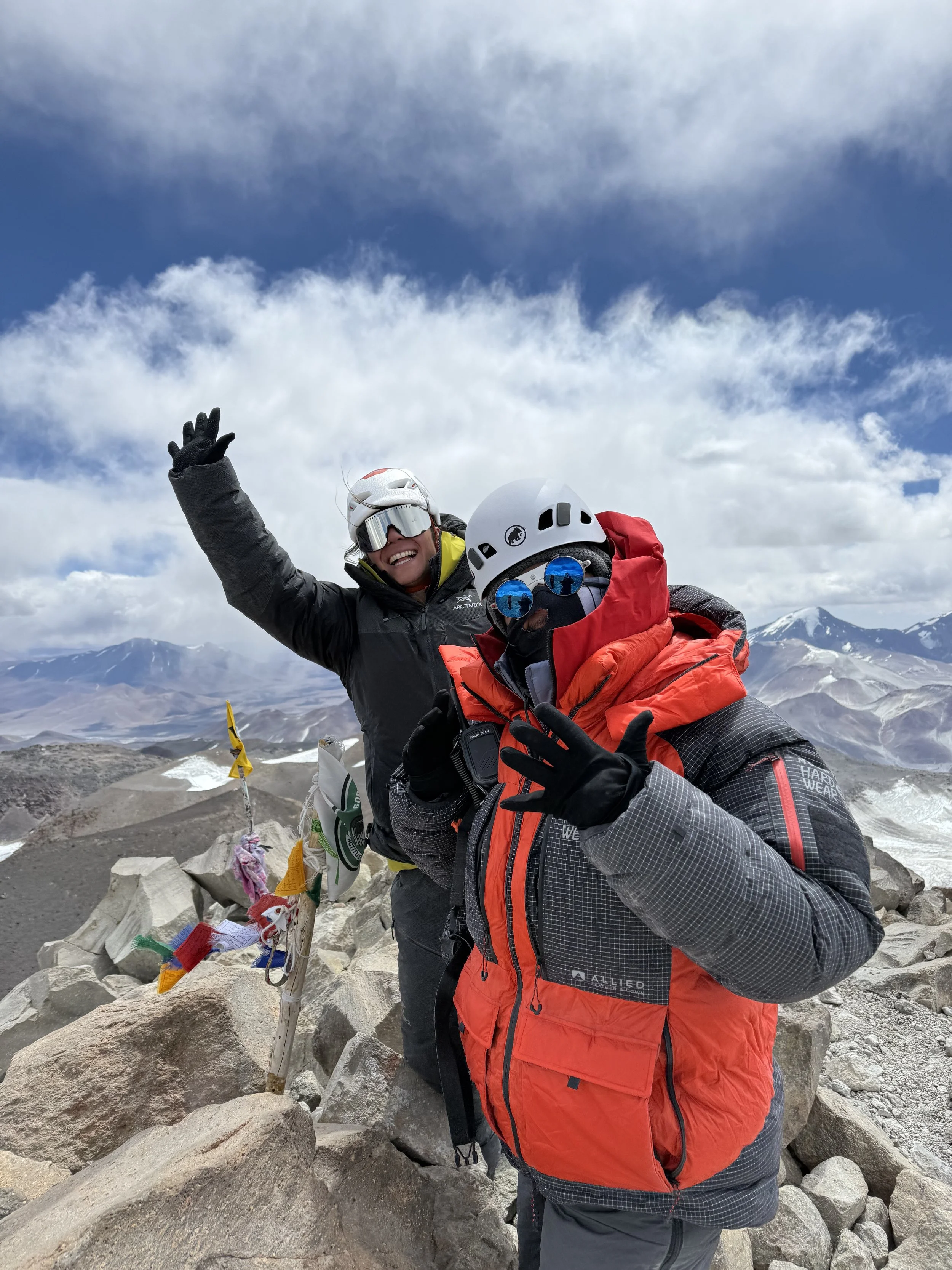Two climbers in winter gear with helmets and sunglasses on a mountain summit, surrounded by rocky terrain and a cloudy sky.